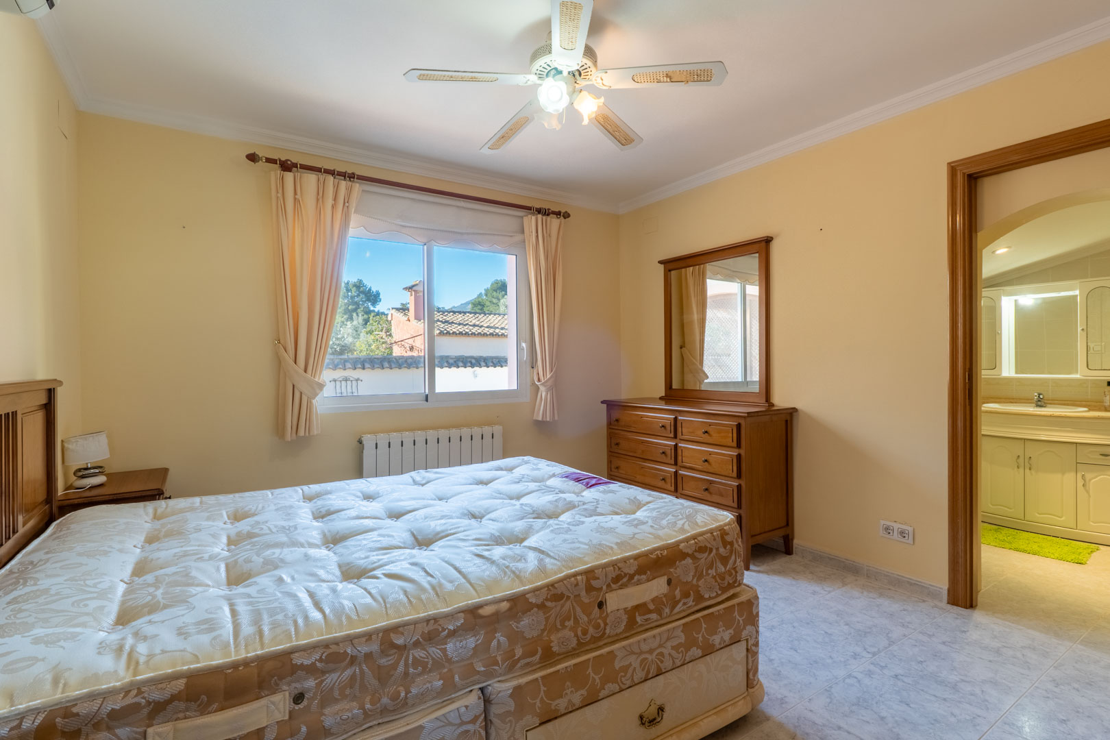 Chambre principale avec mobilier en bois, ventilateur de plafond et salle de bain attenante. La pièce dispose d'un radiateur et d'une fenêtre lumineuse.