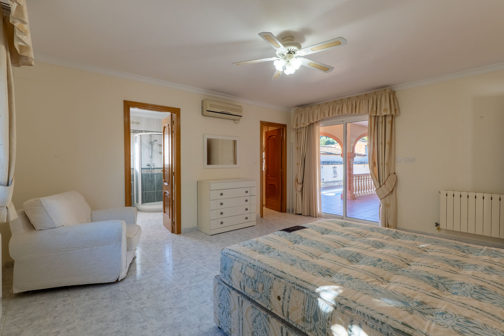 Chambre principale avec salle de bain attenante, climatisation et accès direct à la terrasse. Elle dispose de carrelage clair, d'un ventilateur de plafond et de vues extérieures.