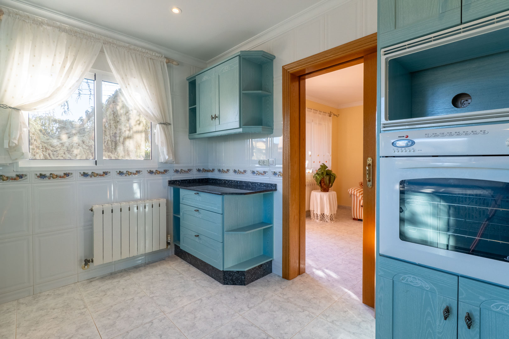 Cuisine indépendante avec des meubles bleu clair, un plan de travail en granit, des murs carrelés et une ouverture en bois vers le séjour.