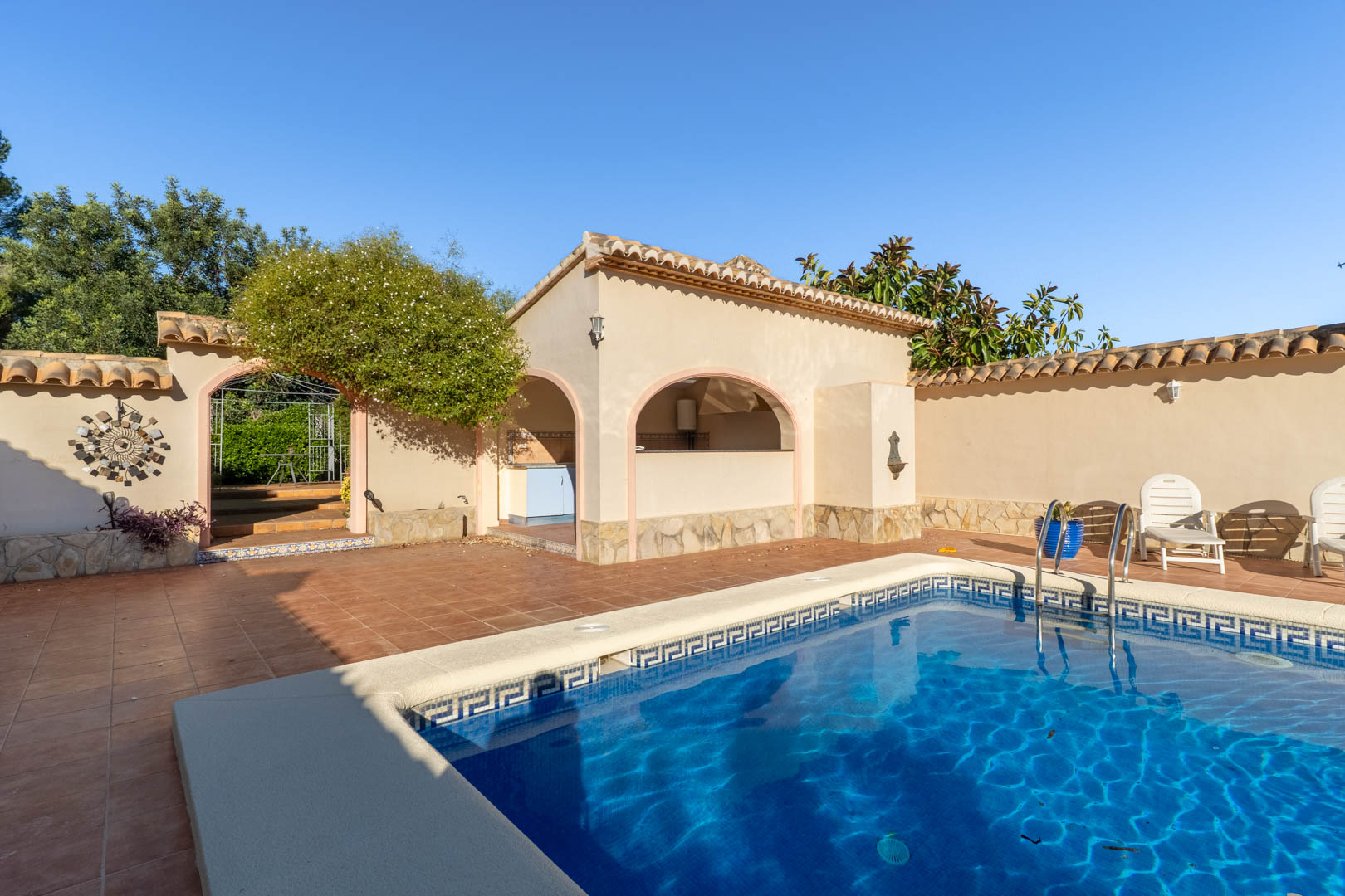 Espace piscine avec terrasse en terre cuite, cuisine d'été abritée par des arches et finitions en pierre naturelle de style méditerranéen.