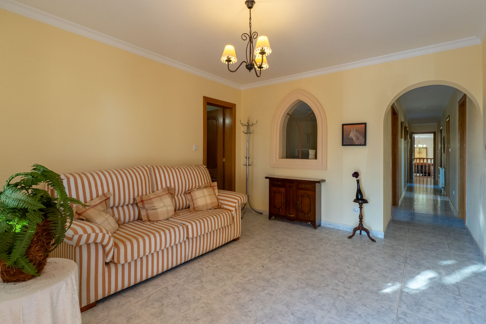 Hall de distribution avec sol en grès cérame, canapé rayé et arche ouvrant sur le couloir. Une fenêtre intérieure décorative en ogive souligne le style.