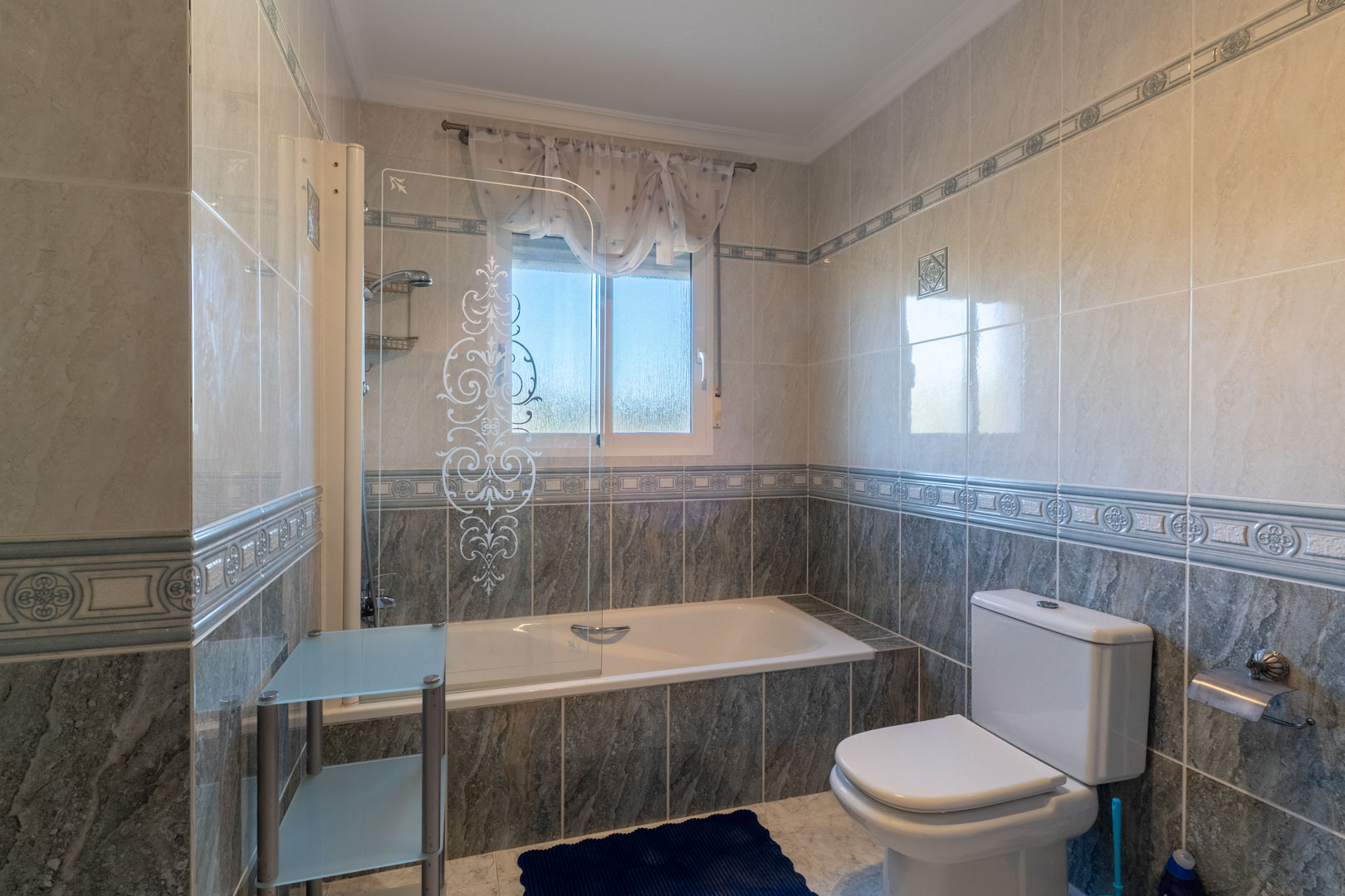 Salle de bain avec baignoire, paroi en verre sérigraphié et carrelage bicolore. Une fenêtre assure une luminosité naturelle et une aération optimale.