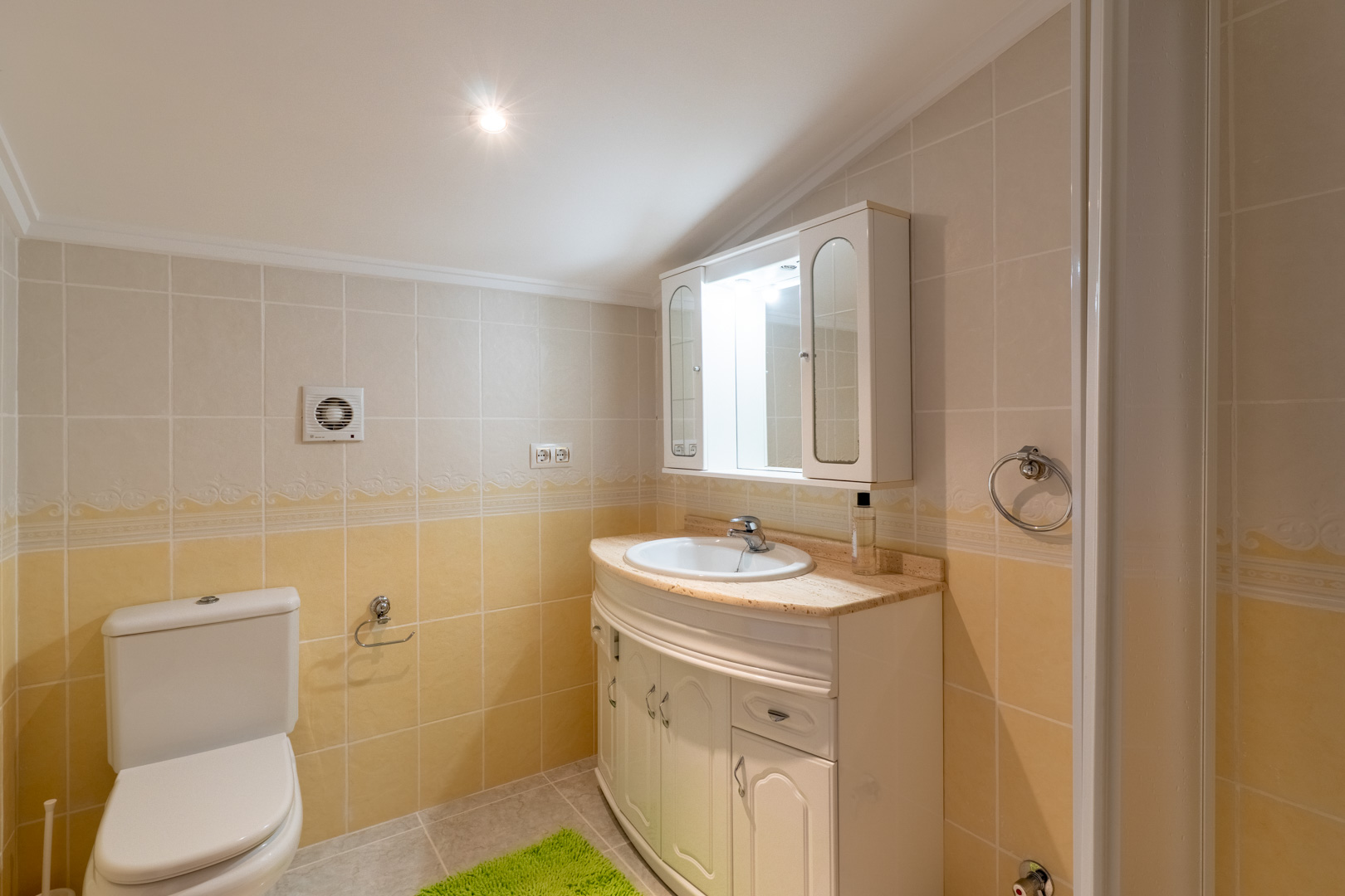 Salle de bain avec carrelage crème et jaune, meuble vasque incurvé avec plan de travail en marbre et armoire de toilette éclairée.