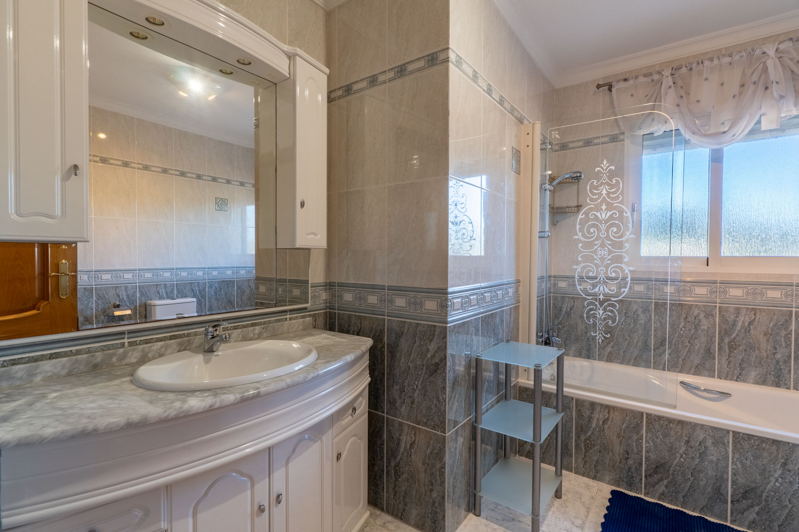 Salle de bain complète avec vasque en marbre, rangements blancs et baignoire avec paroi décorative. Murs carrelés avec frises et fenêtre apportant de la lumière naturelle.