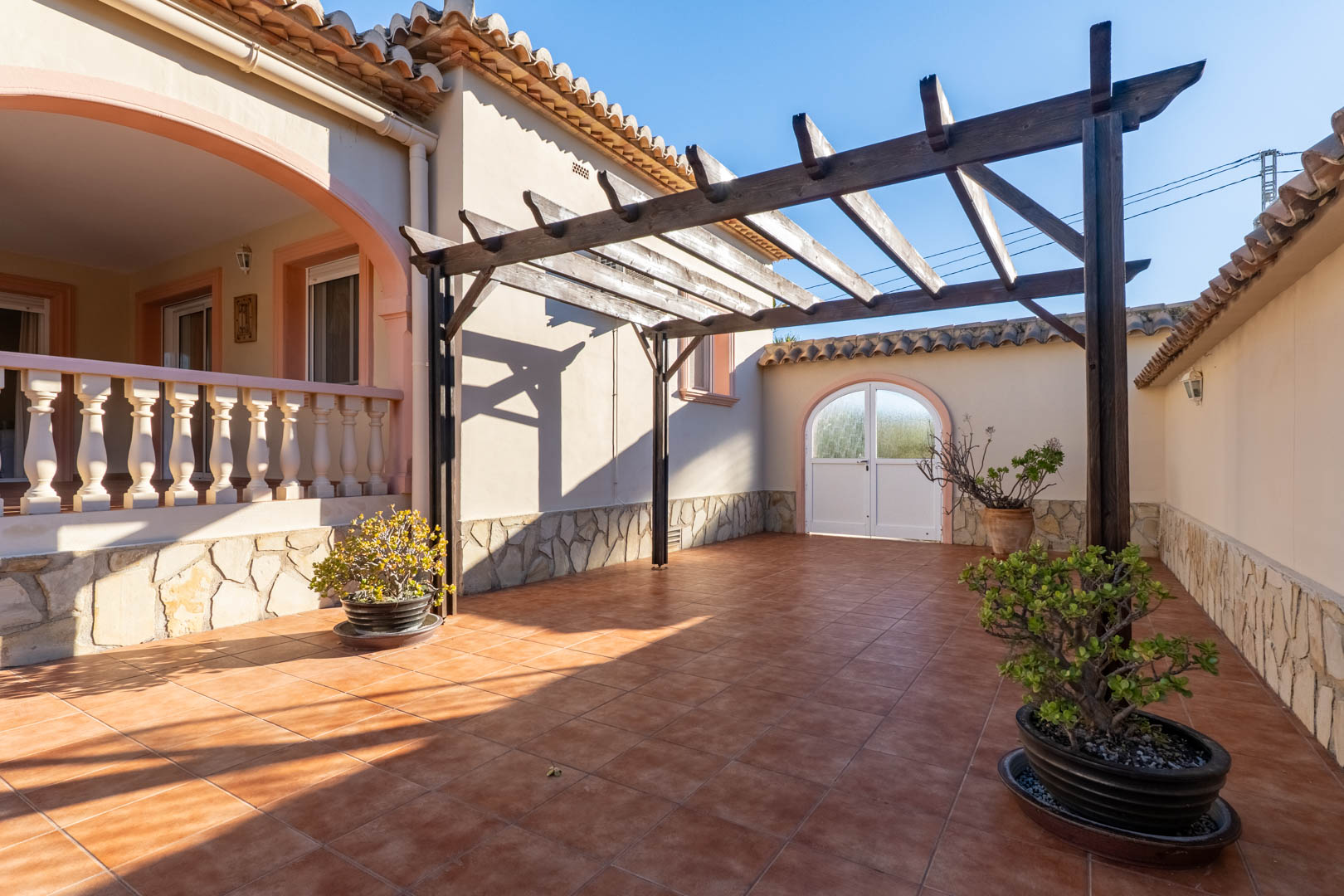 Terrasse extérieure avec carrelage en terre cuite, pergola en bois et soubassement en pierre. Elle communique avec une naya surélevée bordée d'une balustrade.