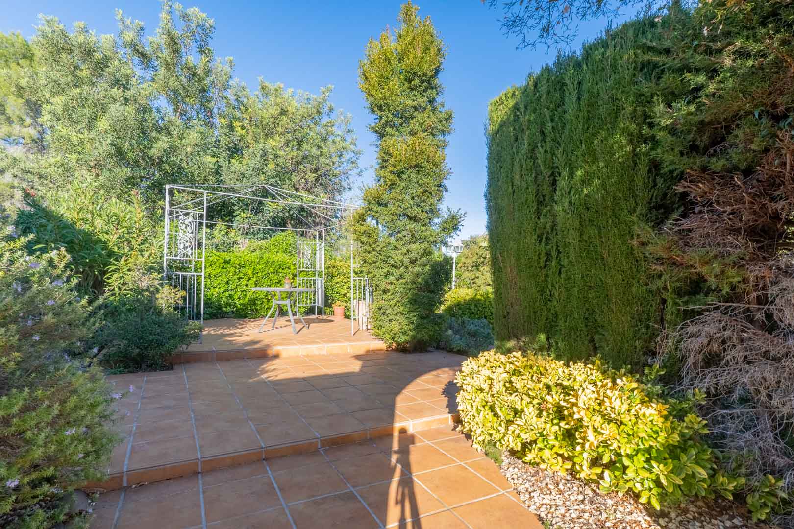 Terrasse extérieure avec sol en grès, pergola métallique blanche et jardin méditerranéen arboré offrant un espace de détente ombragé et privé.