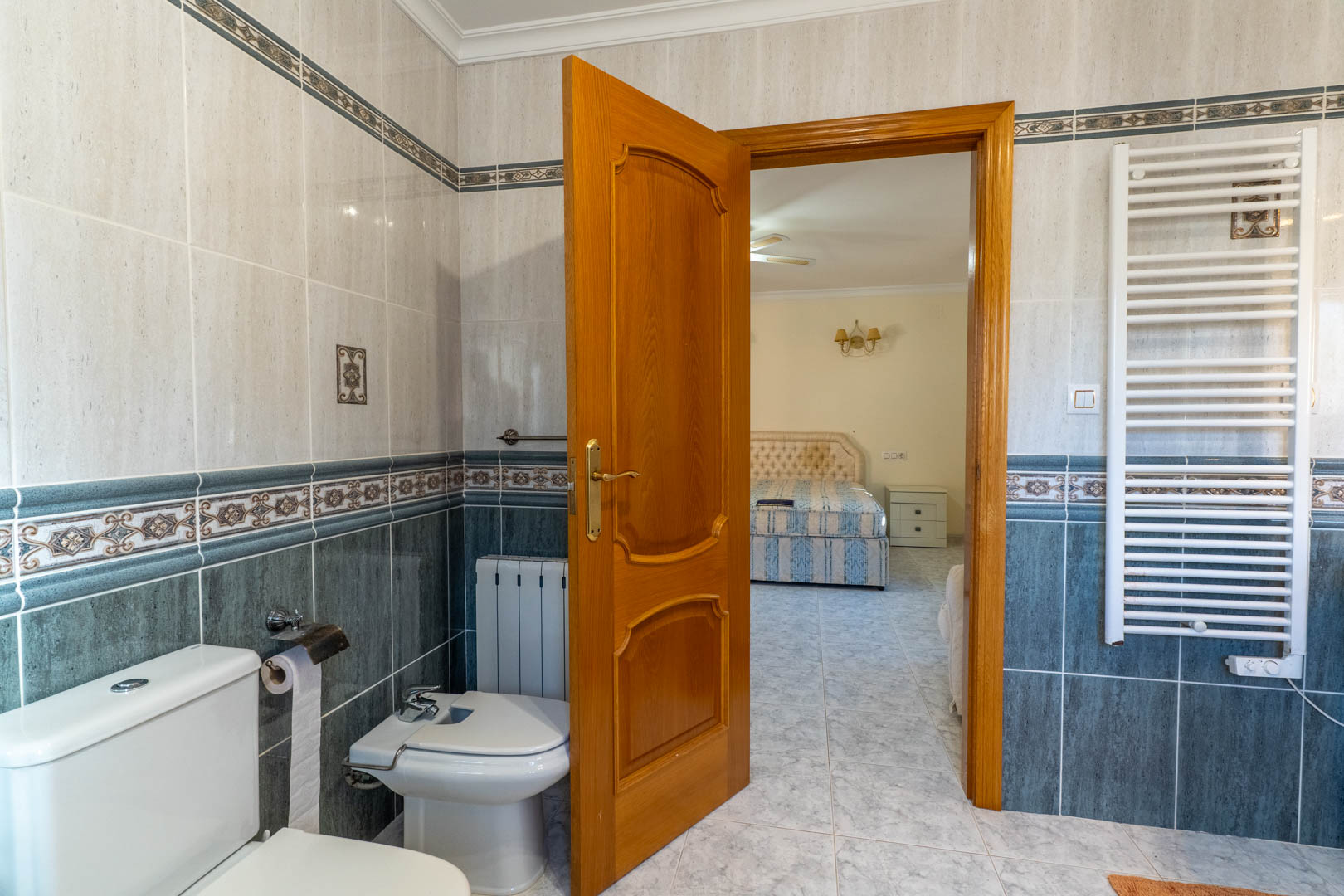 Vue depuis la salle de bain attenante vers la chambre, avec carrelage bicolore, radiateur sèche-serviettes blanc et menuiseries en bois massif.