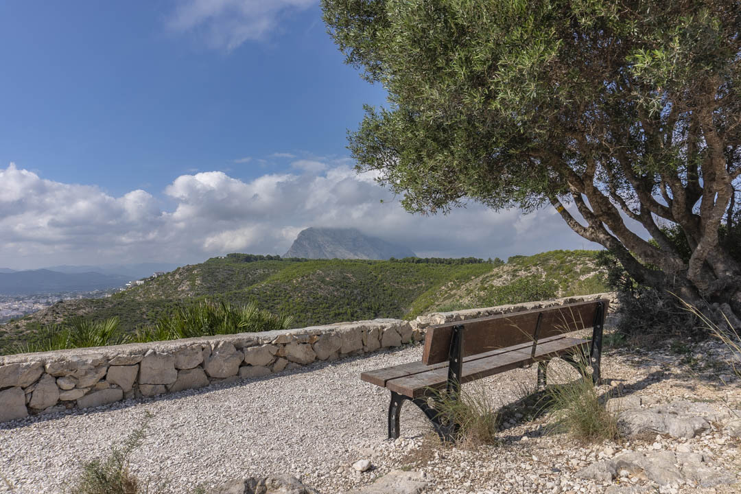 Belvédère avec banc en bois et muret en pierre sèche, offrant une vue panoramique sur le massif du Montgó et les collines verdoyantes.
