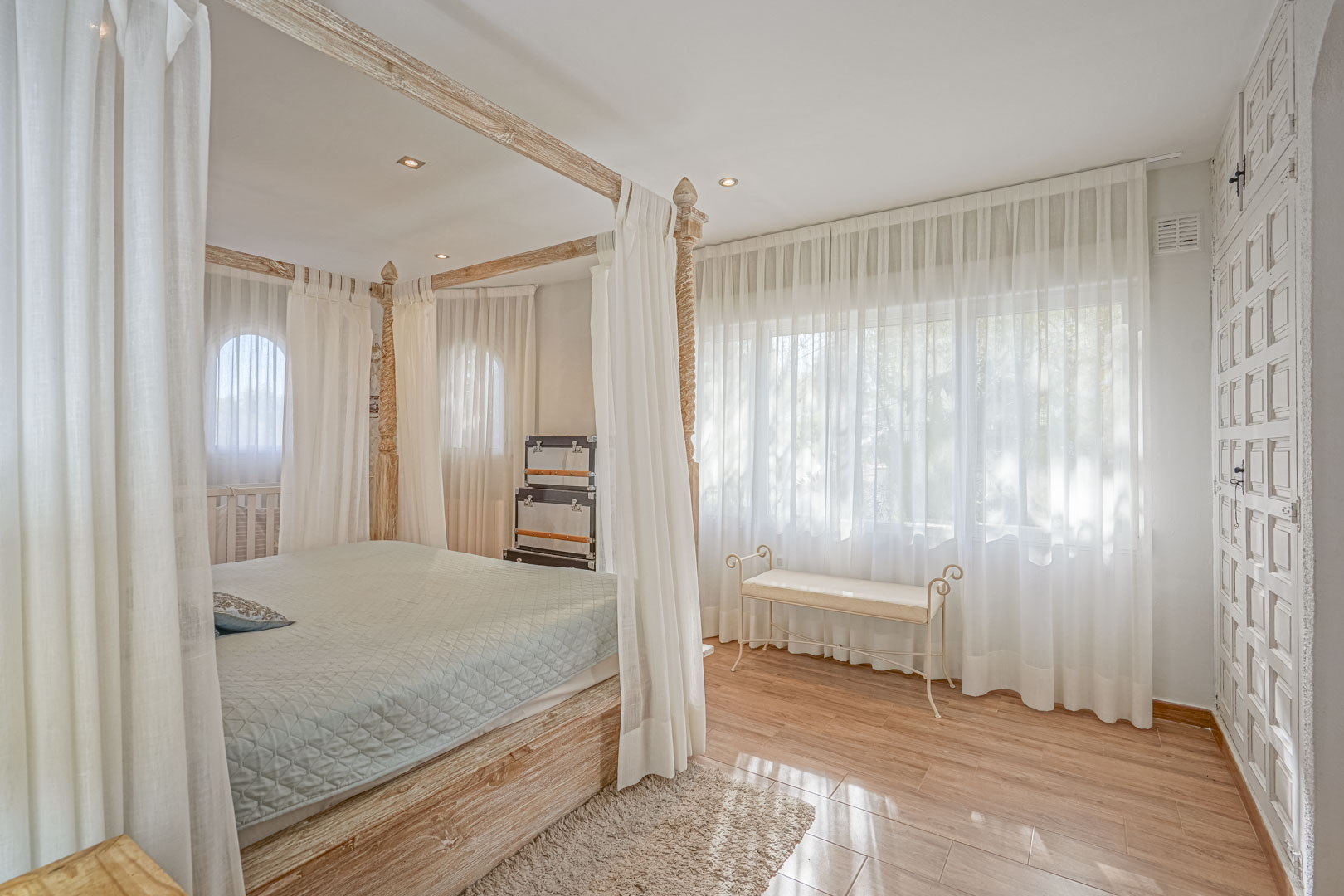 Chambre avec lit à baldaquin en bois clair, sol en céramique imitation bois et grandes fenêtres avec voilages laissant passer une lumière naturelle douce.