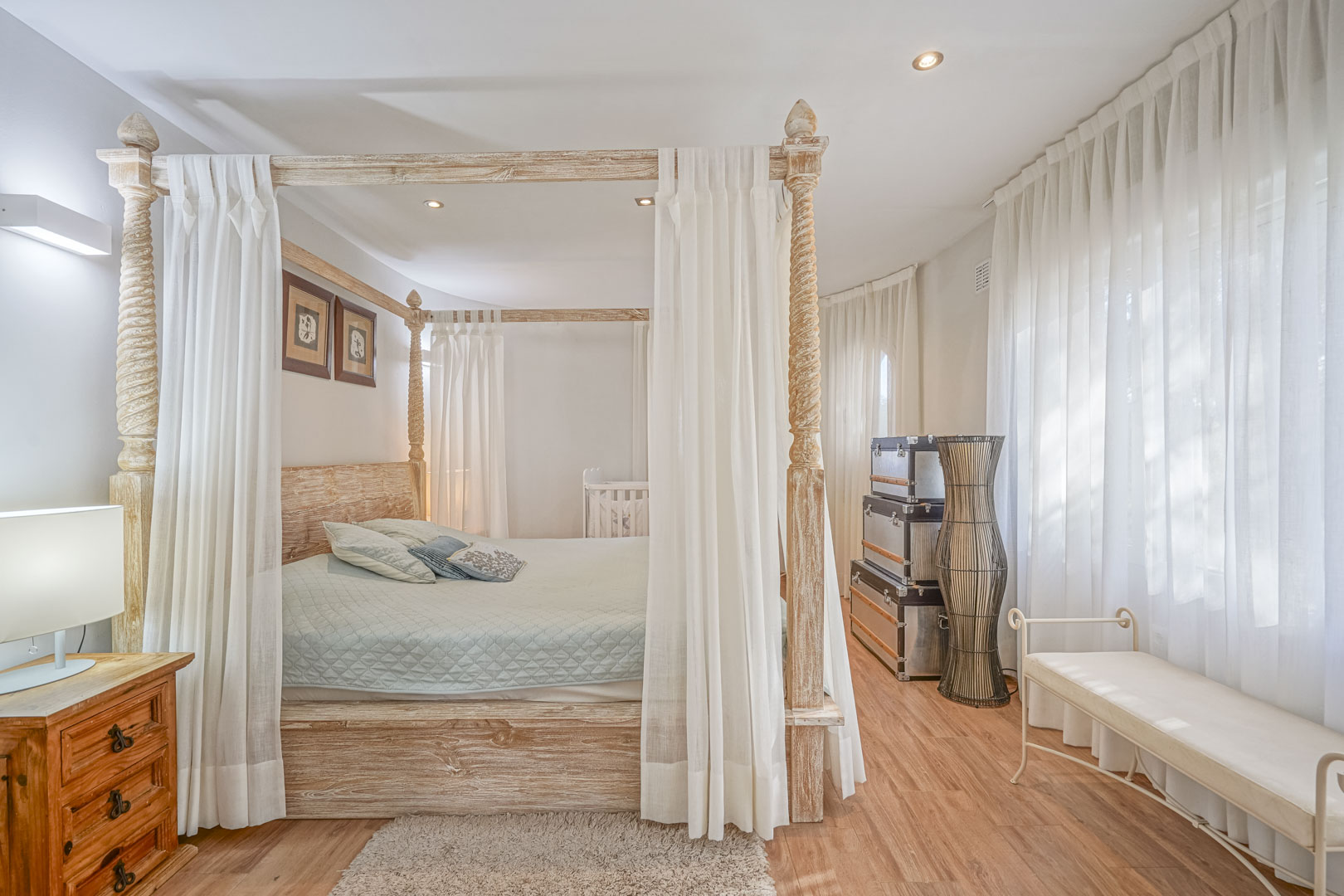 Chambre avec lit à baldaquin en bois sculpté, parquet stratifié et grandes fenêtres parées de voilages blancs pour une lumière naturelle.