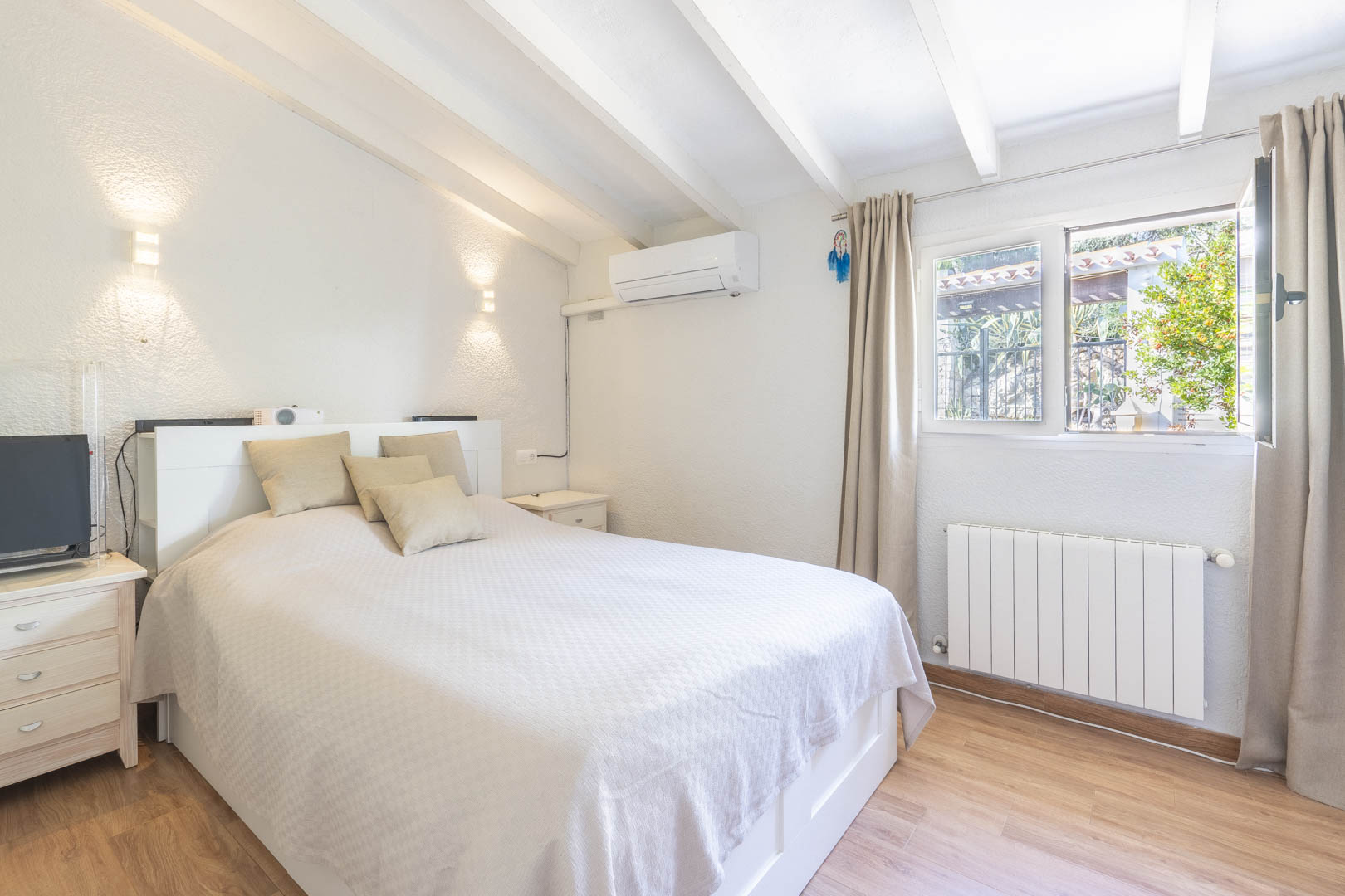 Chambre avec plafond à poutres apparentes blanches, climatisation et radiateur. Elle offre un sol stratifié et une fenêtre lumineuse.