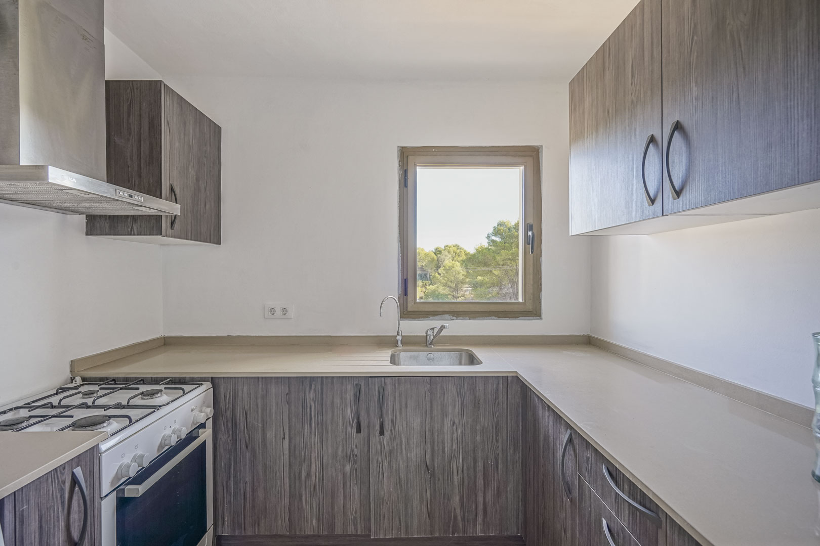 Cuisine fonctionnelle avec placards effet bois sombre, plan de travail neutre et fenêtre vue sur nature. Équipée d'une cuisinière à gaz et d'une hotte en acier inoxydable.