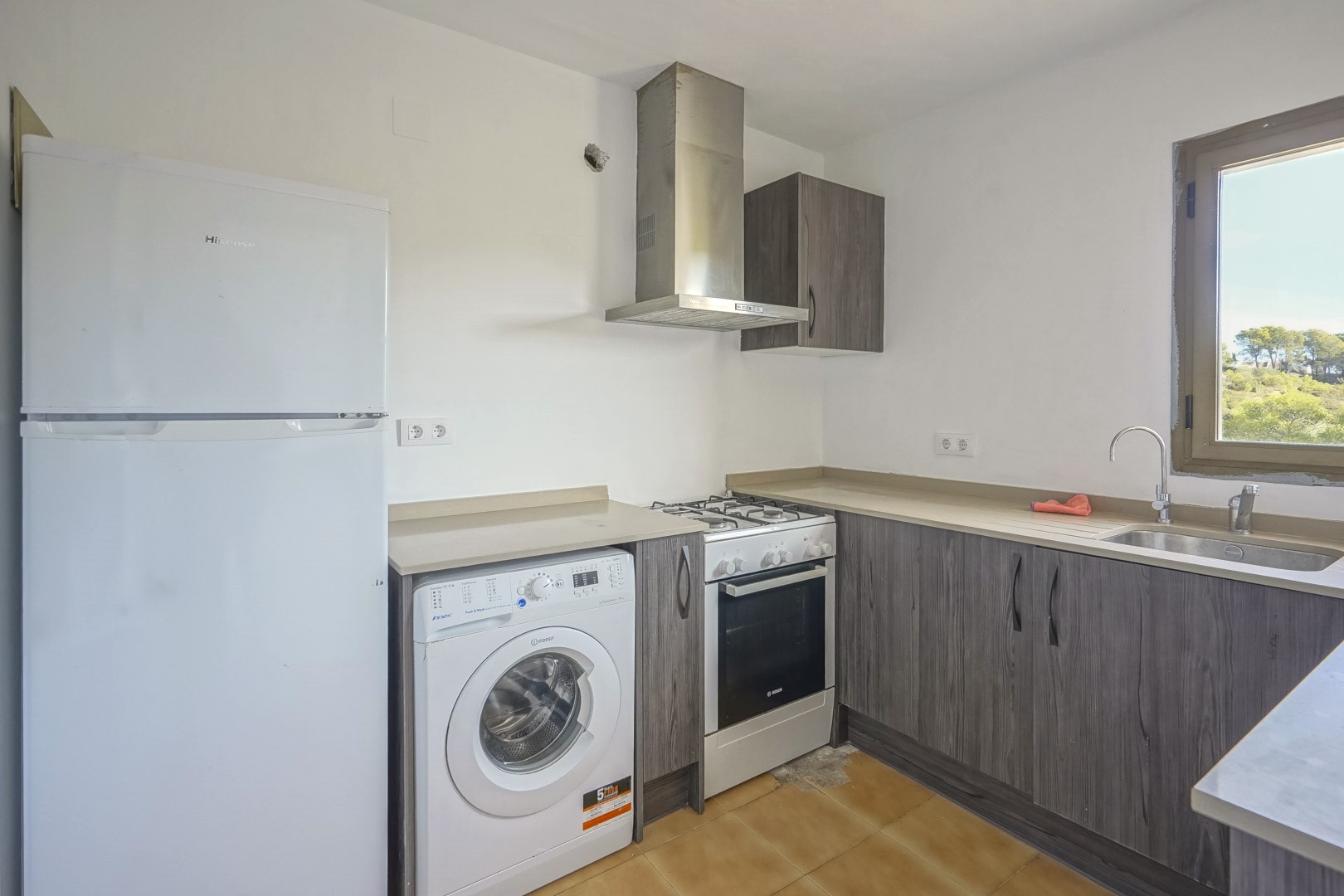 Cuisine fonctionnelle avec placards gris effet bois, équipée d'un four à gaz, d'un lave-linge et d'une fenêtre offrant une vue sur la végétation.