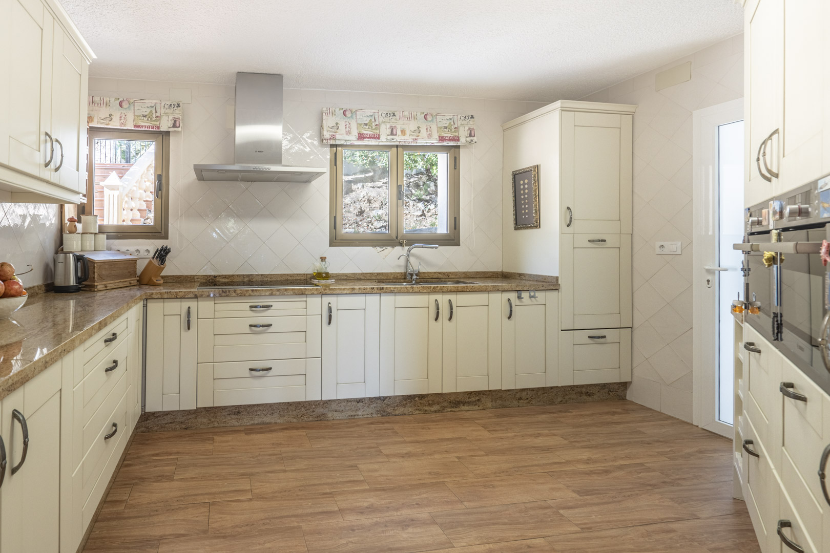 Cuisine rénovée avec placards de couleur crème, plans de travail en granit, hotte en inox et carrelage au sol imitation bois sous une lumière naturelle.