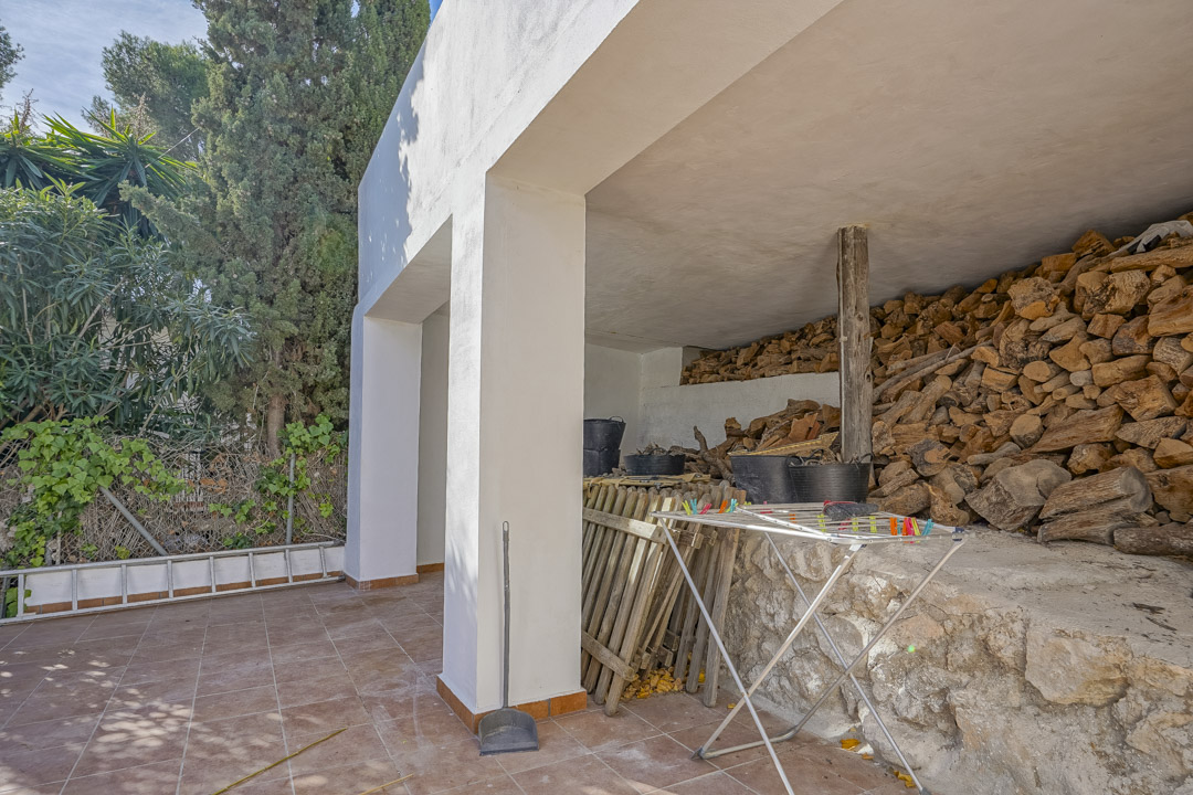 Espace de rangement extérieur couvert avec terrasse carrelée, piliers blancs et une réserve de bois de chauffage sur un soubassement en pierre.