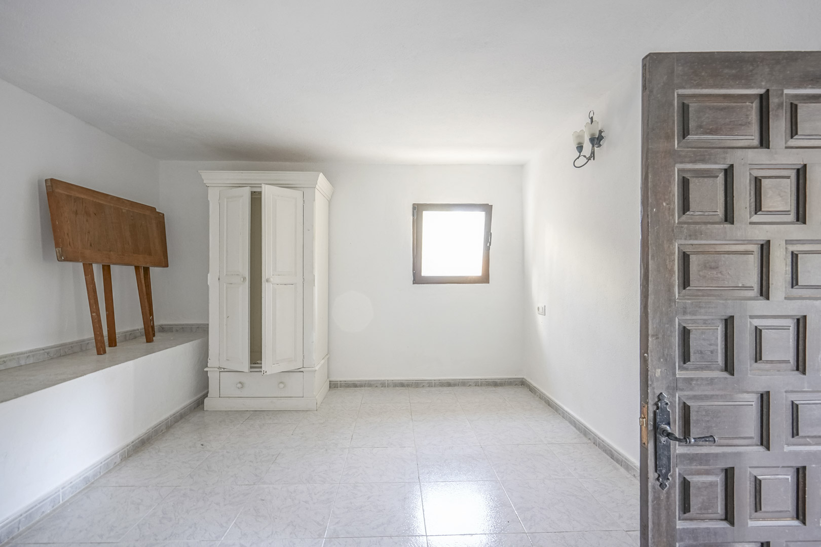 Pièce lumineuse avec sol en carrelage clair, murs blancs et porte en bois sculpté. Elle dispose d'une armoire blanche classique et d'une petite fenêtre.