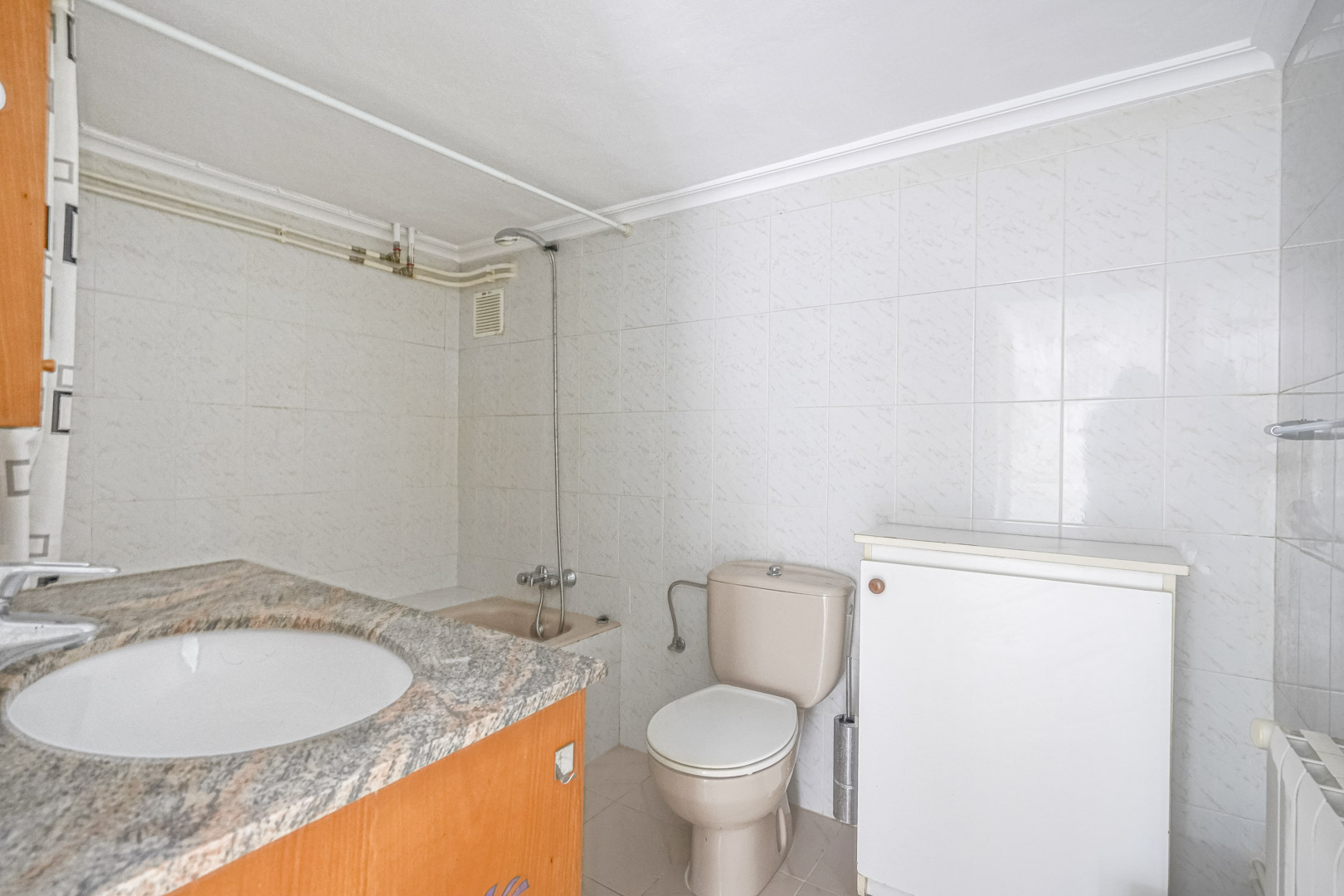 Salle de bain avec plan de travail en granit, lavabo encastré, toilettes beiges et murs carrelés en blanc. Comprend un espace douche et un radiateur mural.
