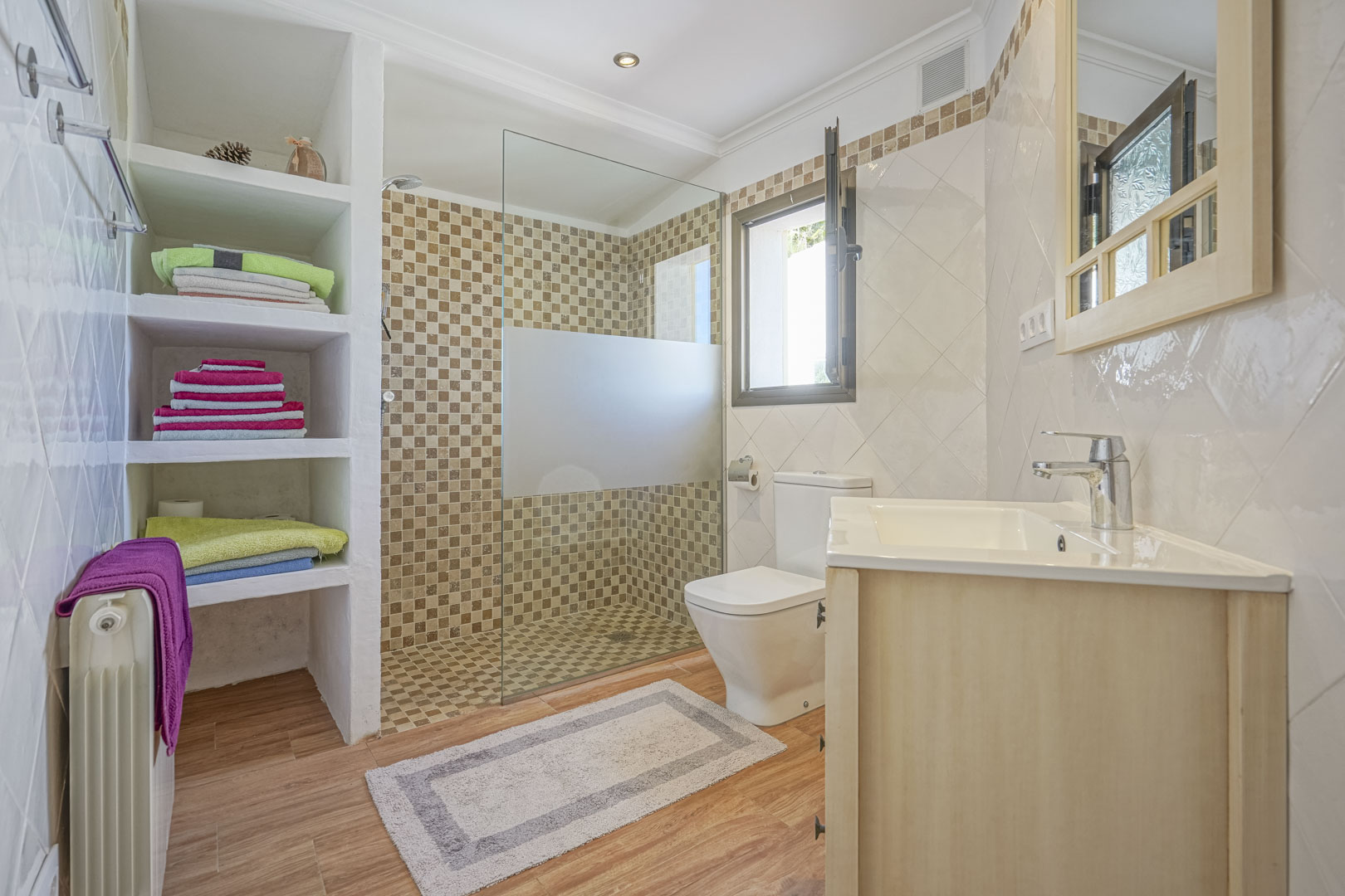 Salle de bain rénovée avec douche à l'italienne en mosaïque, étagères intégrées en maçonnerie, sol imitation bois et meuble vasque en bois clair.