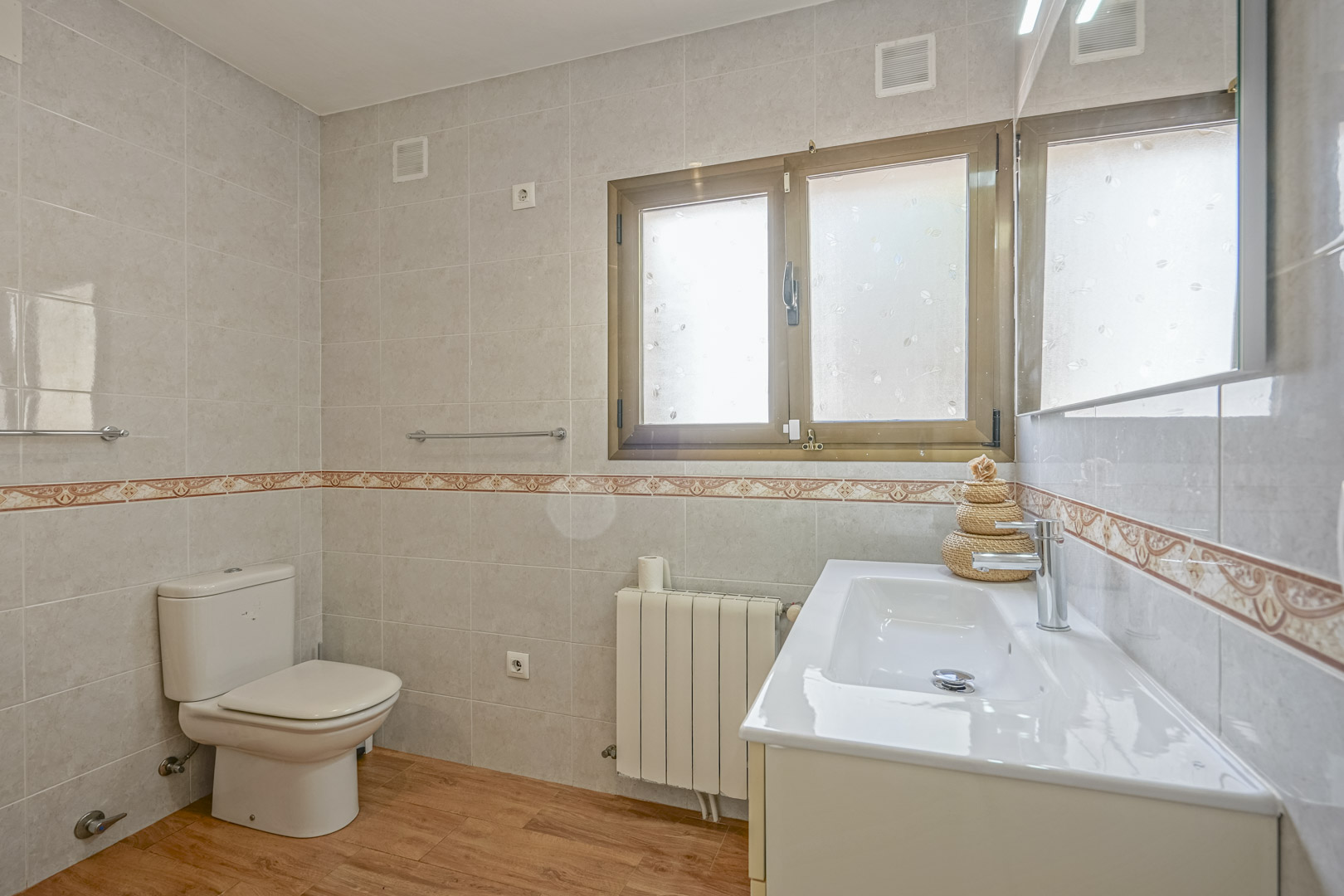 Salle de bain rénovée avec sol imitation bois, murs carrelés avec frise décorative, meuble vasque blanc et radiateur sous une fenêtre dépolie.