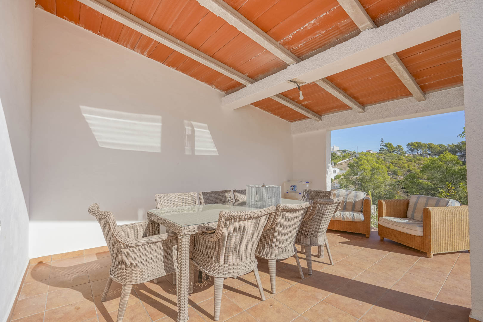Terrasse couverte avec plafond à poutres et briques, comprenant un coin repas en osier et offrant une vue dégagée sur les collines environnantes.