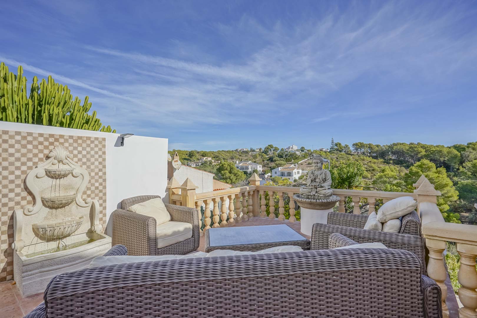 Terrasse surélevée avec mobilier en rotin, fontaine murale décorative et balustrade en pierre offrant une vue dégagée sur les collines environnantes.