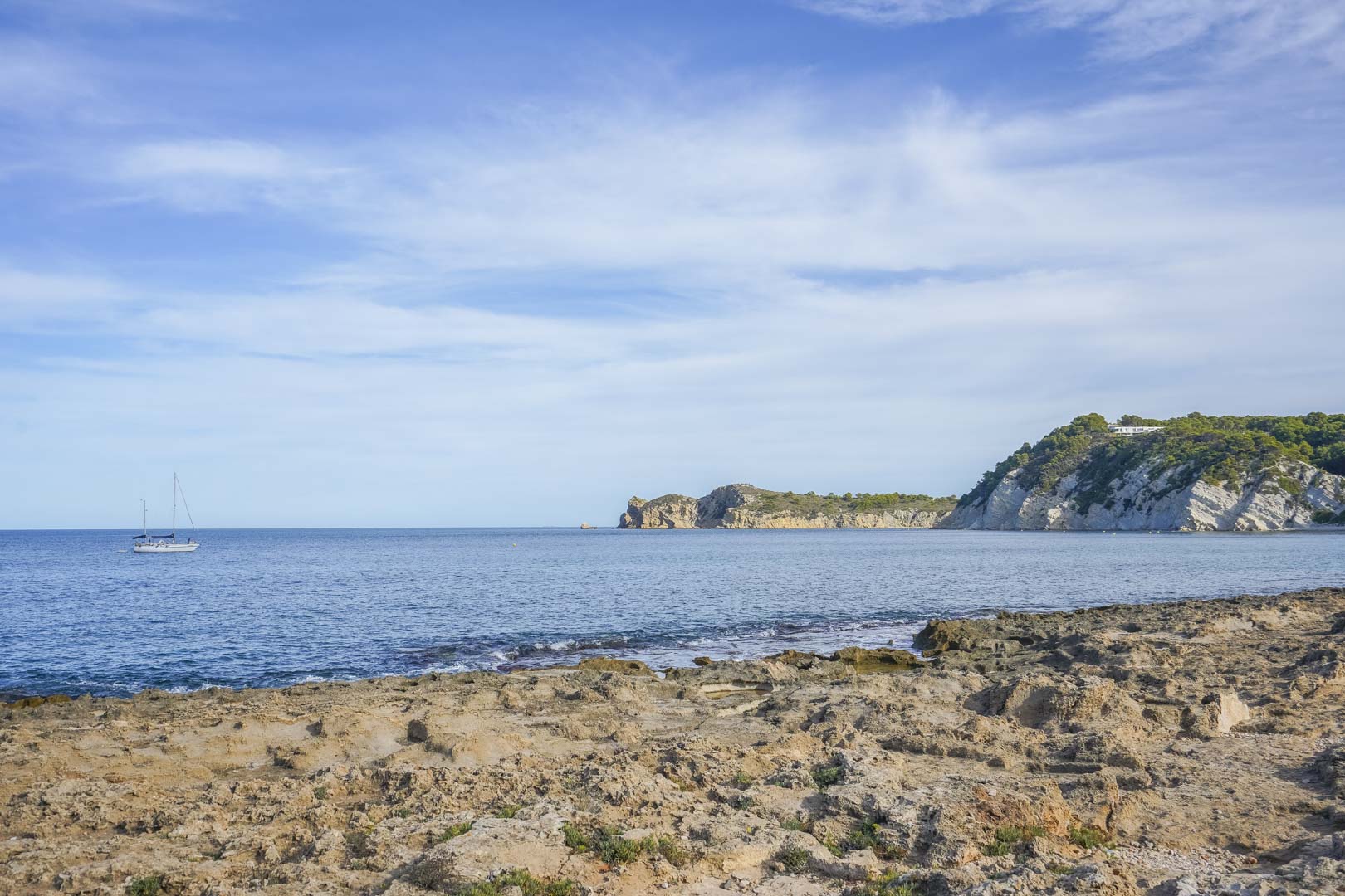 Vue de la côte rocheuse de Jávea avec la mer Méditerranée, des falaises de calcaire et un voilier naviguant sous un ciel dégagé.