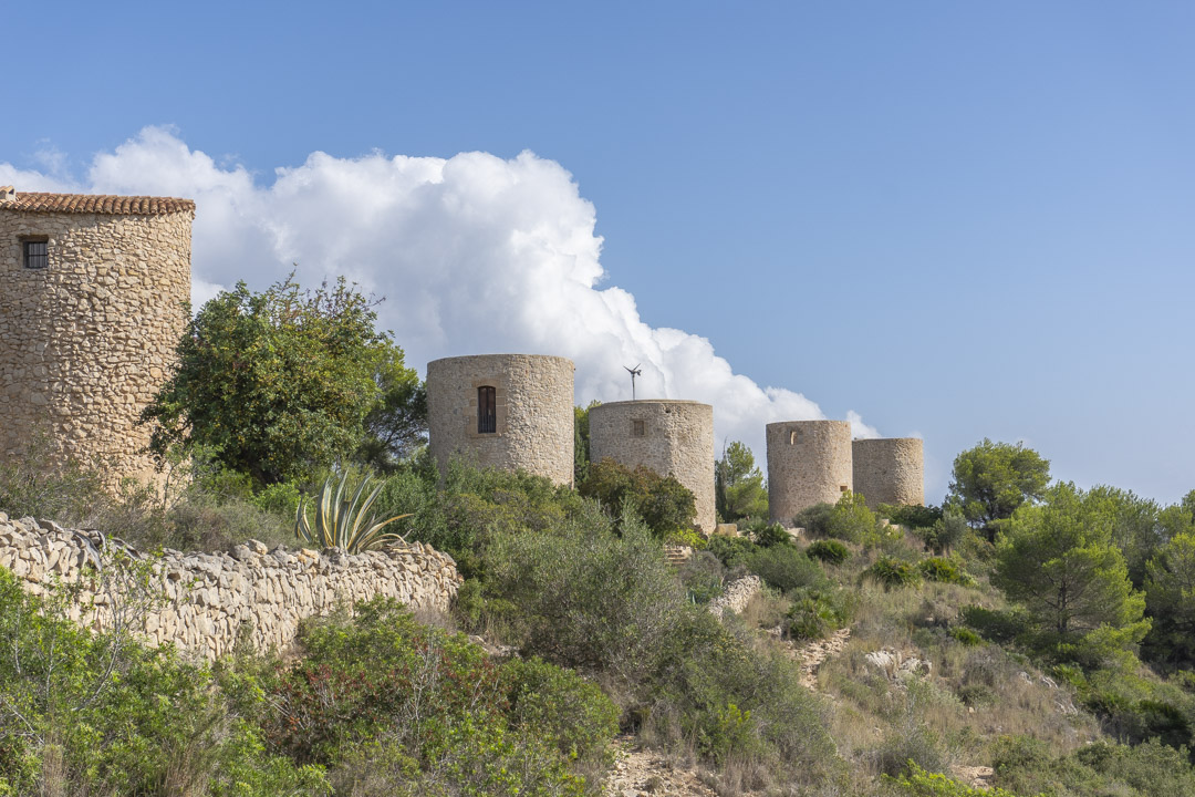 Vue des moulins à vent historiques en pierre de Jávea, situés sur une colline avec une végétation méditerranéenne et des murs en pierre sèche.
