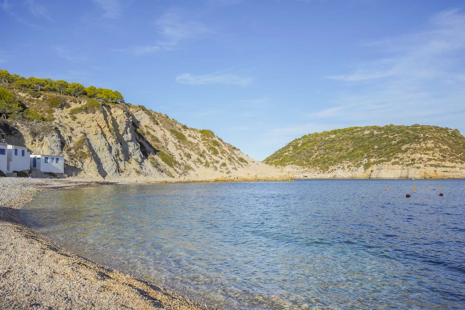 Vue d'une crique de galets à Jávea aux eaux cristallines, entourée de falaises rocheuses et de végétation méditerranéenne sous un ciel bleu.