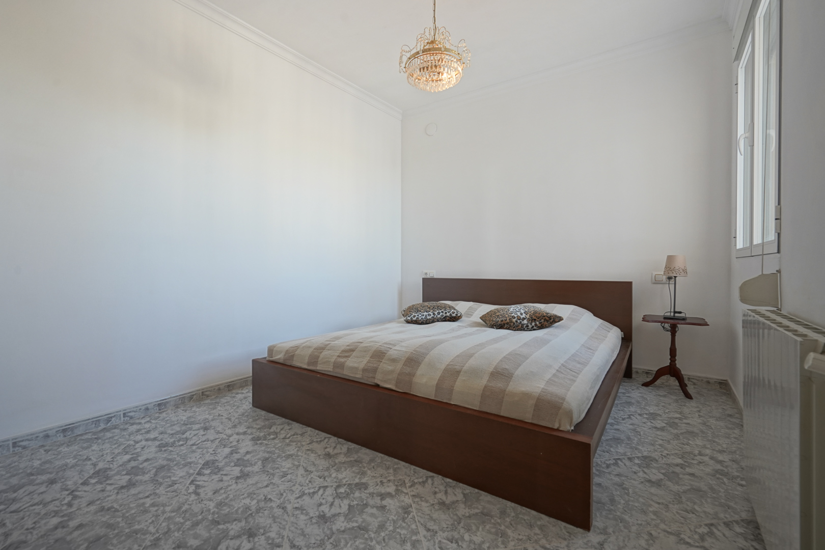 Chambre avec cadre de lit en bois sombre, sol en grès cérame marbré, lustre classique et radiateur installé sous la fenêtre.