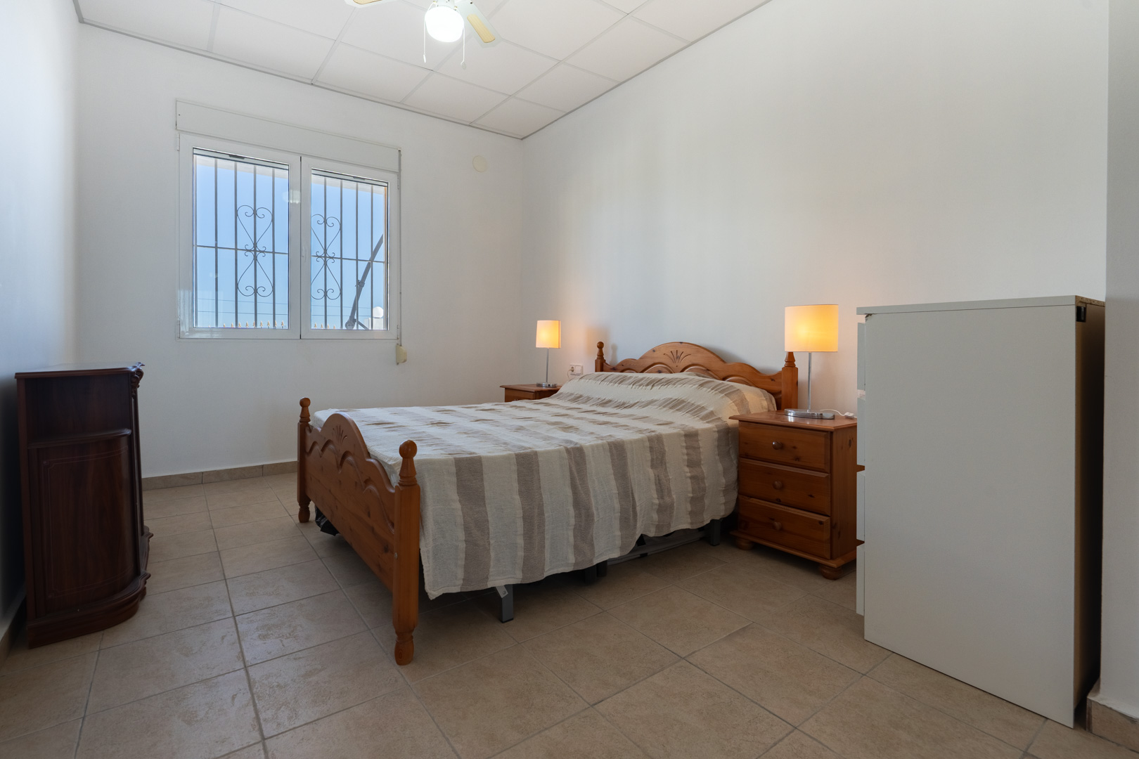Chambre avec mobilier en bois traditionnel, sol en grès et fenêtre équipée de volets roulants et de grilles de sécurité offrant une lumière naturelle.