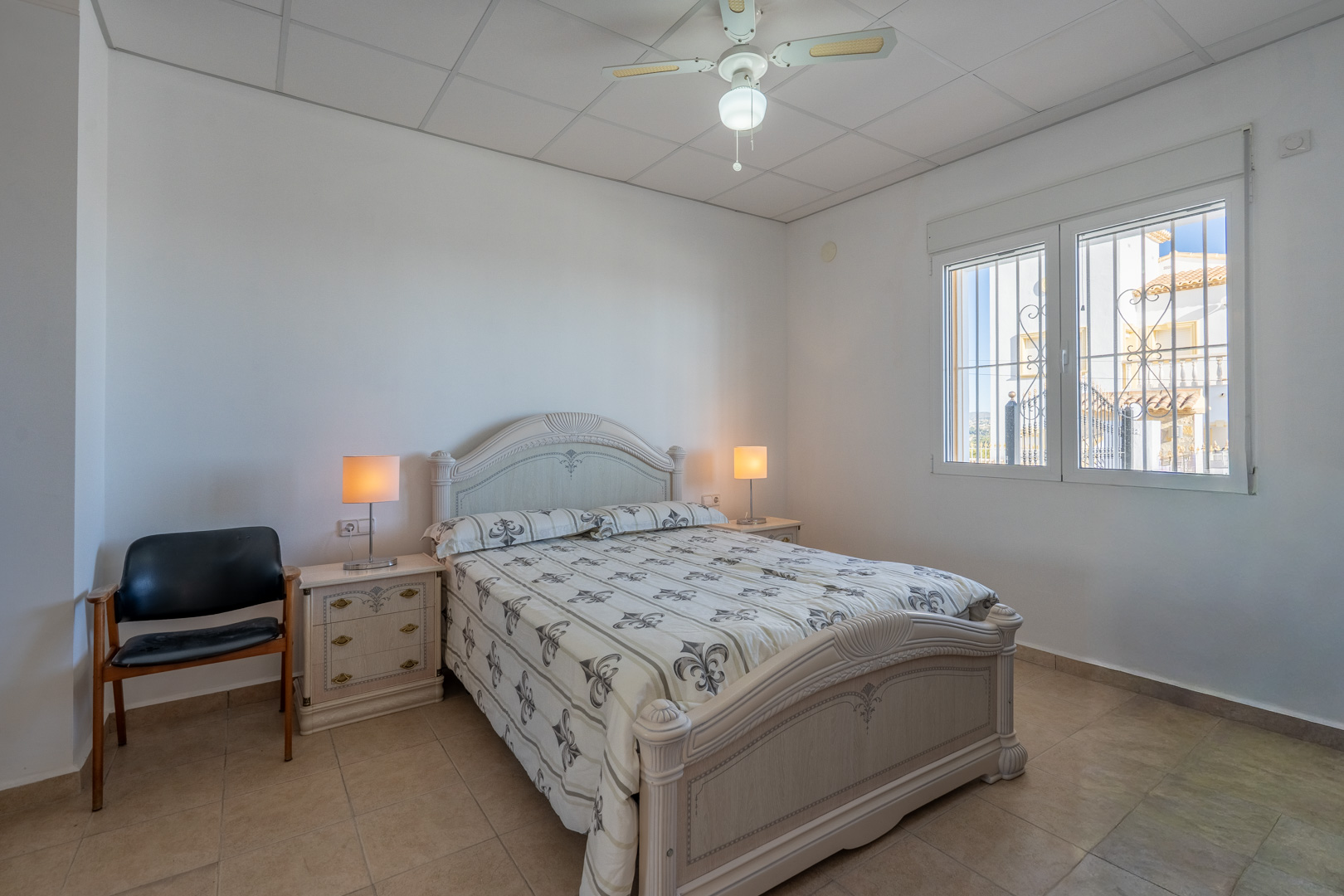 Chambre avec mobilier en bois traditionnel, sol en grès, ventilateur de plafond et fenêtre avec grilles de sécurité offrant une lumière naturelle.