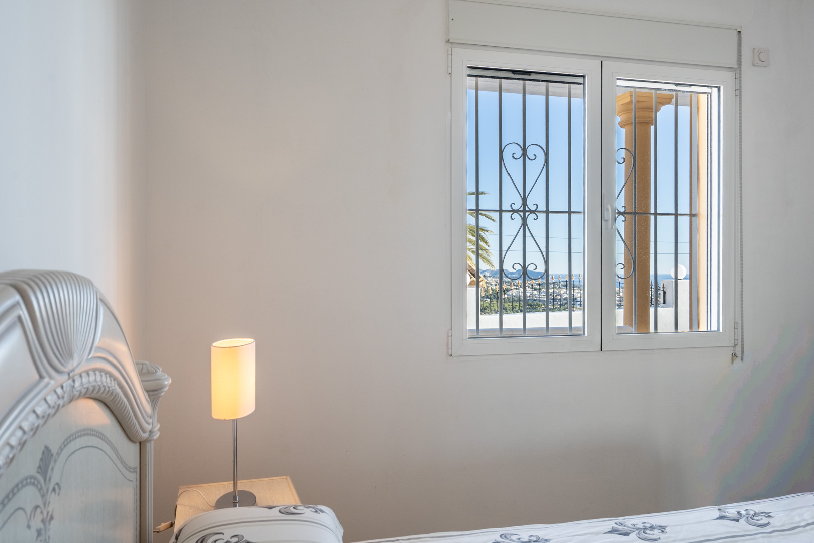 Chambre avec tête de lit sculptée et fenêtre aux grilles ornementales offrant une vue sur le littoral de Calpe et la mer Méditerranée.