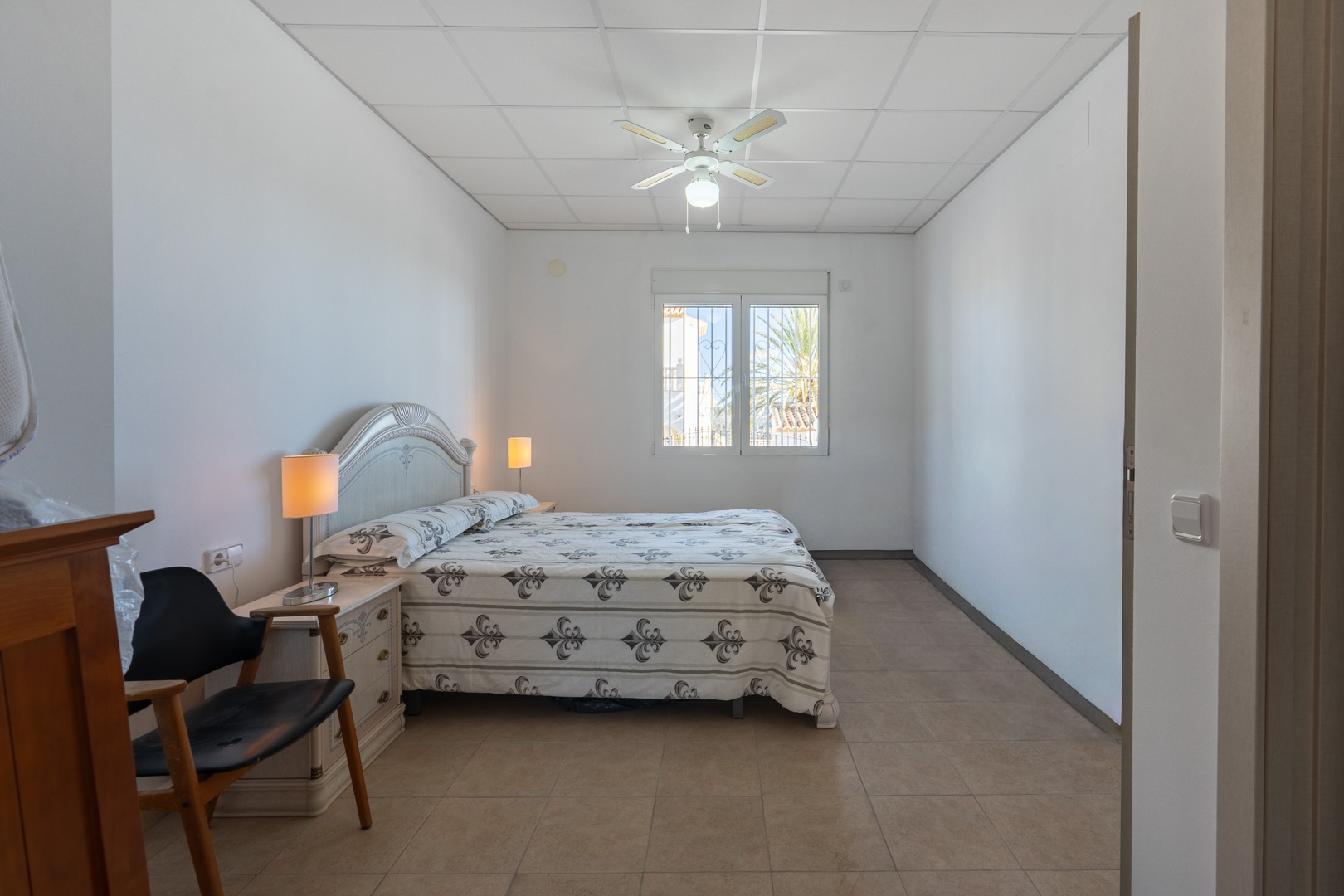 Chambre lumineuse avec lit double, ventilateur de plafond et sol en grès. Une fenêtre apporte de la lumière naturelle et offre une vue sur la végétation extérieure.