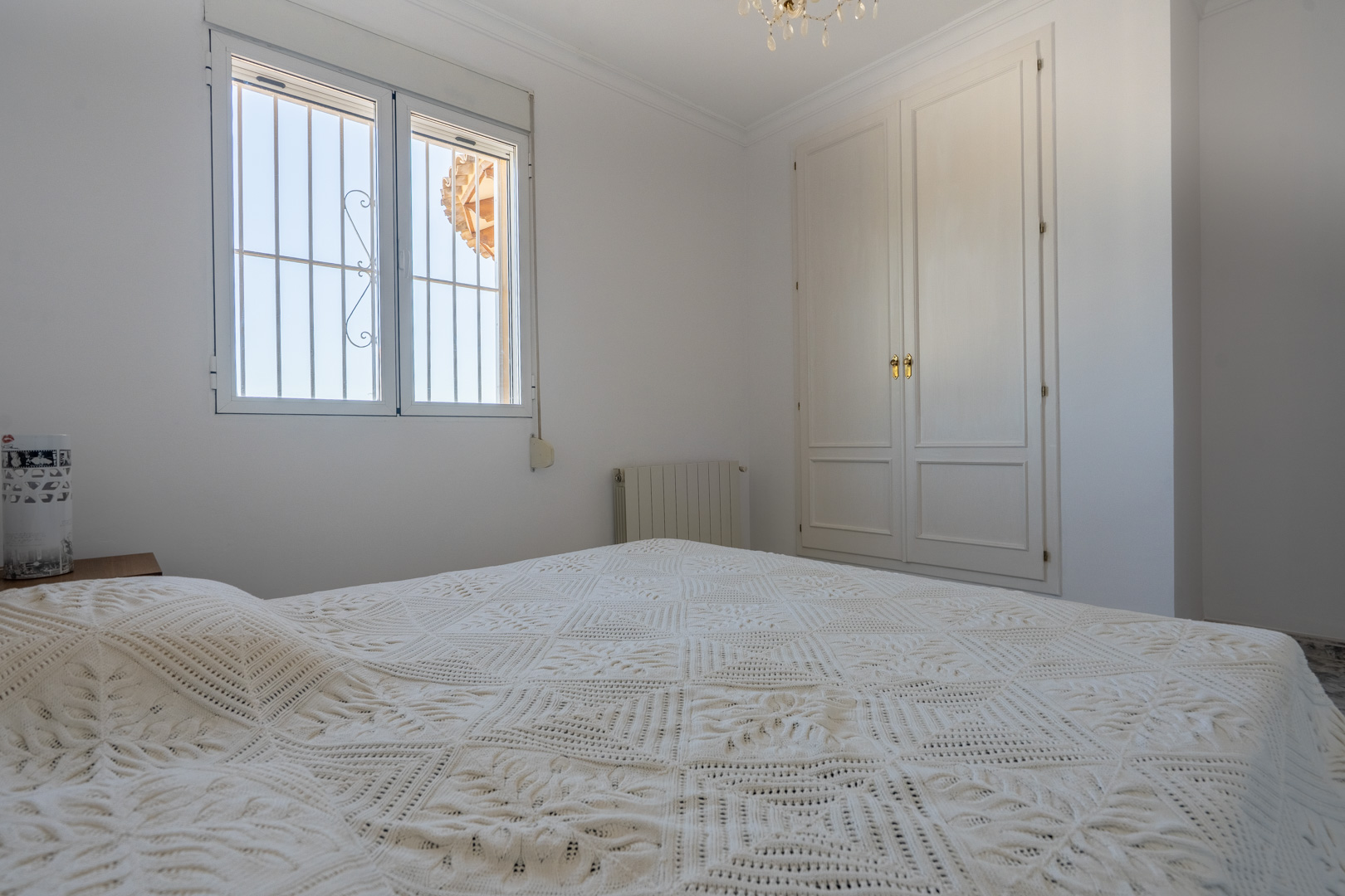 Chambre lumineuse avec murs blancs, armoire encastrée en bois laqué et fenêtre avec grilles de sécurité. Équipée d'un radiateur et bénéficiant d'un éclairage naturel.