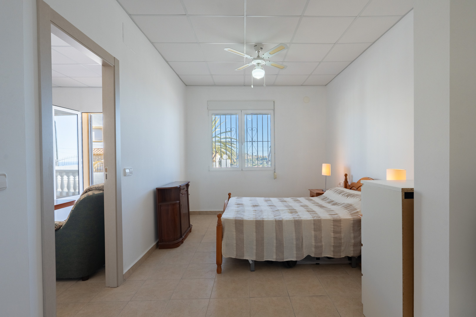 Chambre lumineuse avec sol en grès cérame, ventilateur de plafond et fenêtre sécurisée. La pièce communique avec le séjour et offre une vue sur la végétation extérieure.