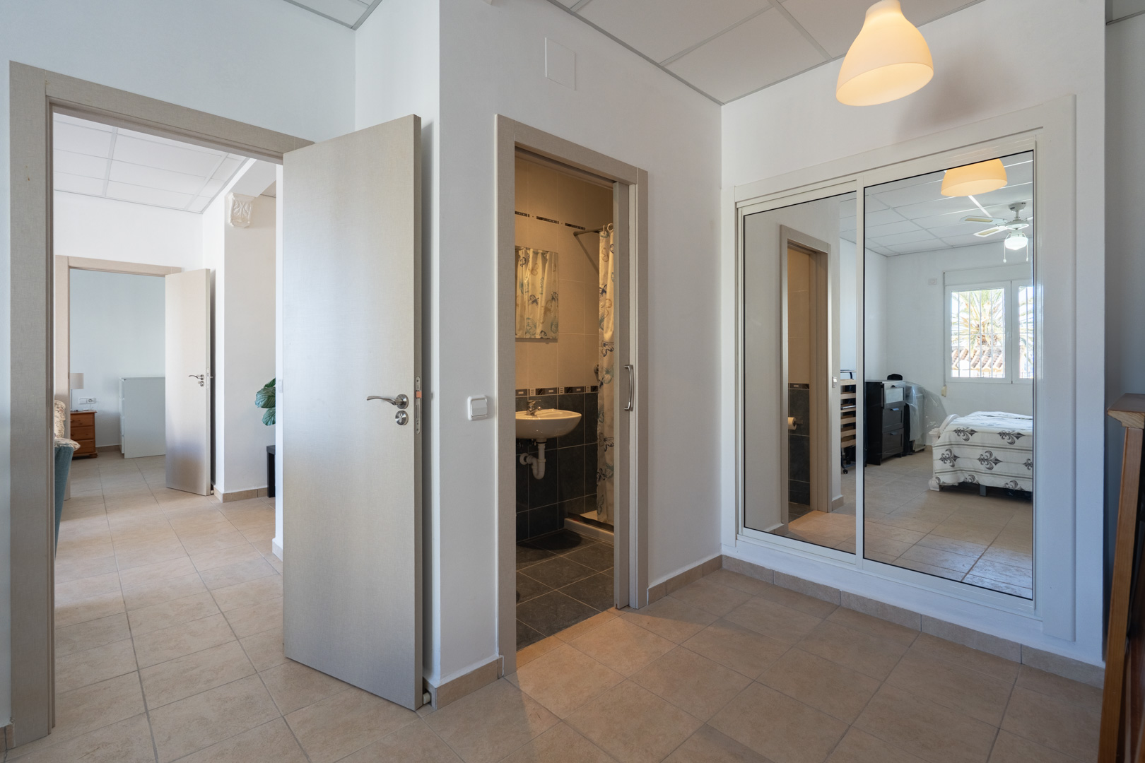 Couloir de distribution avec sol en grès, placard intégré à portes miroirs et accès à une salle de bain et une chambre équipée d'un ventilateur de plafond.