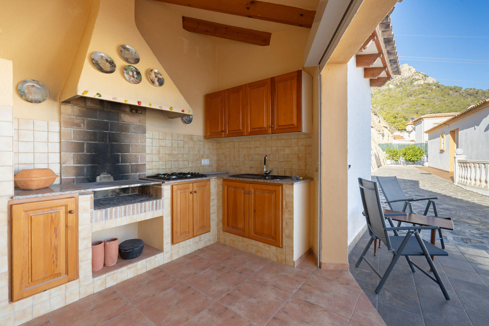Cuisine d'été couverte avec barbecue en brique, meubles en bois, plan de travail en granit et évier, donnant sur une terrasse avec vue sur la Sierra de Oltà.