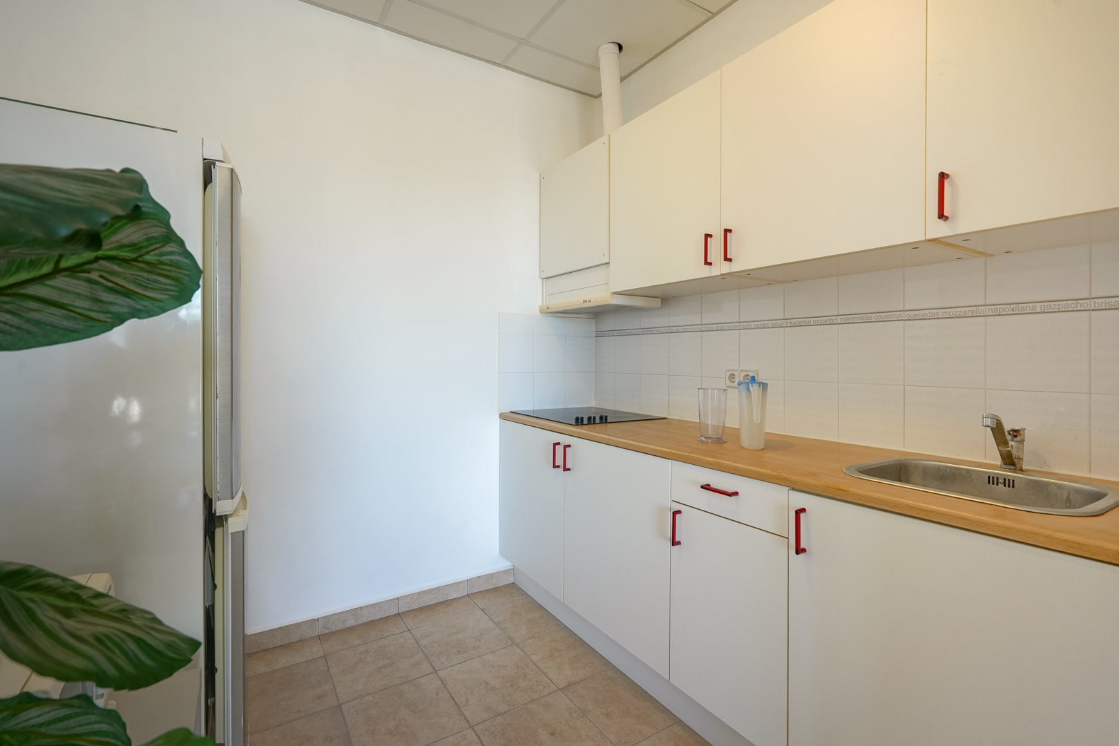 Cuisine fonctionnelle avec placards blancs, poignées rouges, plan de travail en bois et crédence carrelée. Équipée d'une plaque de cuisson et d'un évier intégré.