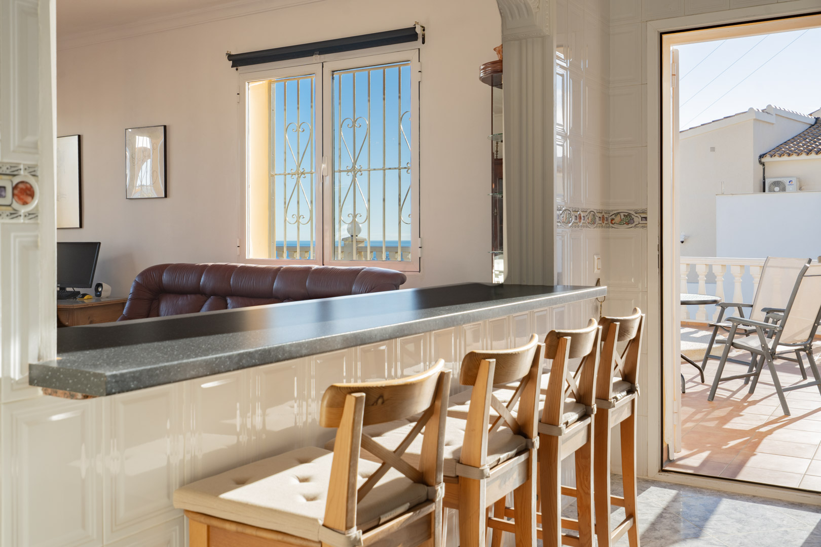 Cuisine semi-ouverte avec bar de petit-déjeuner sombre et tabourets en bois, s'ouvrant sur le salon et une terrasse ensoleillée avec vue sur mer.