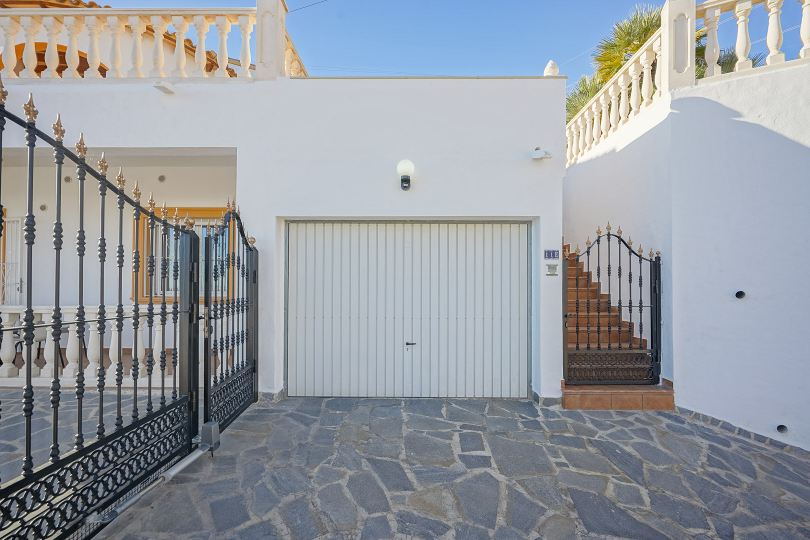 Entrée de la propriété avec garage fermé, portail en fer forgé et escalier extérieur, le tout sur un revêtement en pierre naturelle.
