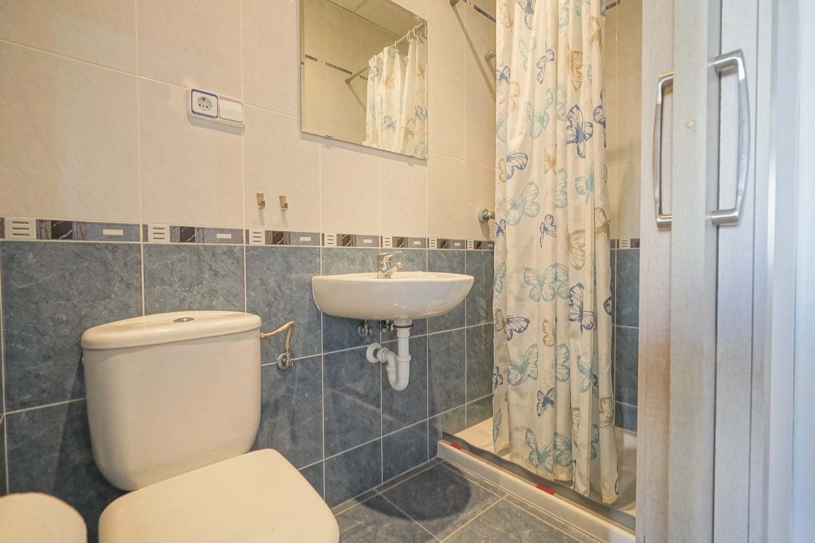 Salle de bain avec carrelage bicolore bleu et crème, comprenant un lavabo suspendu, des toilettes et un espace douche avec rideau à motifs.