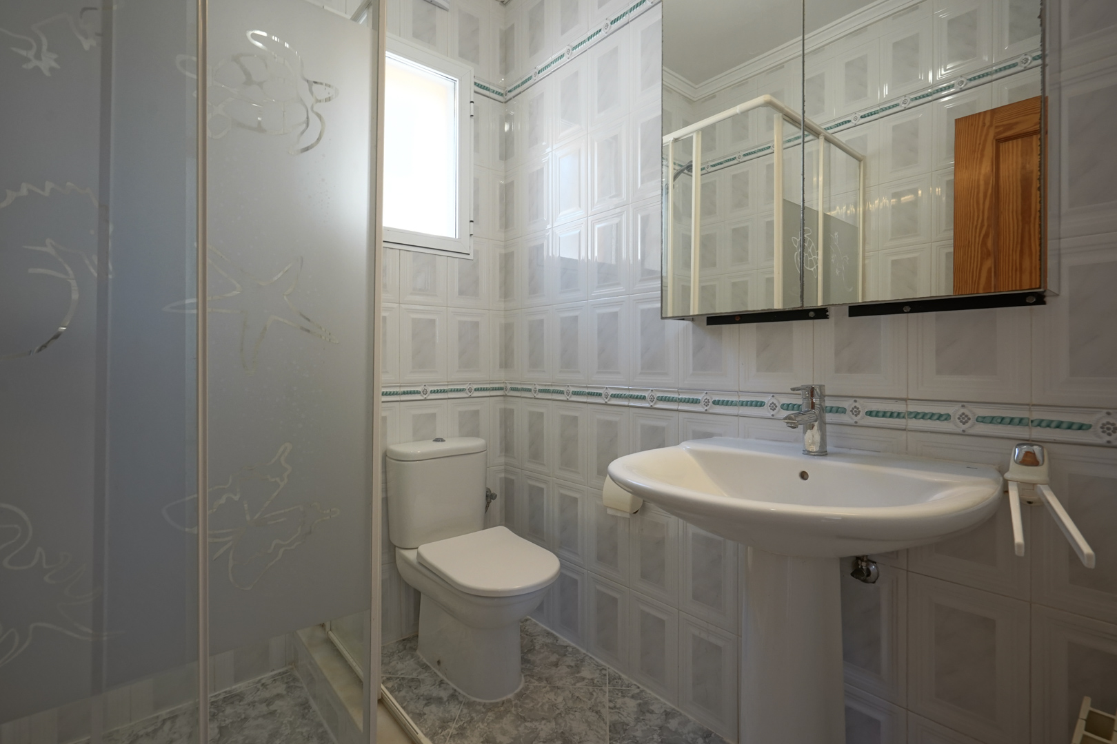 Salle de bain équipée d'un lavabo sur colonne, d'un WC et d'une douche avec paroi en verre dépoli. Les murs sont revêtus de carrelage avec une frise décorative.