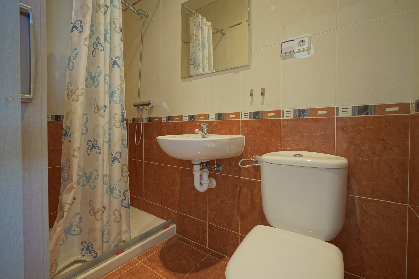 Salle de bain fonctionnelle avec douche, rideau à motifs papillons, lavabo mural et carrelage bicolore dans les tons terre cuite et crème.
