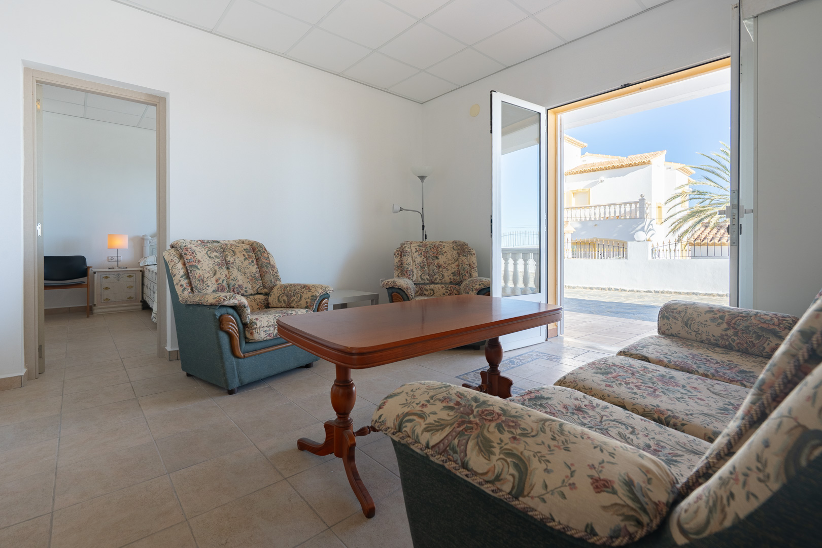 Séjour de l'appartement d'invités avec canapés à motifs floraux, table en bois et accès terrasse. On aperçoit une chambre attenante et un sol en grès cérame.