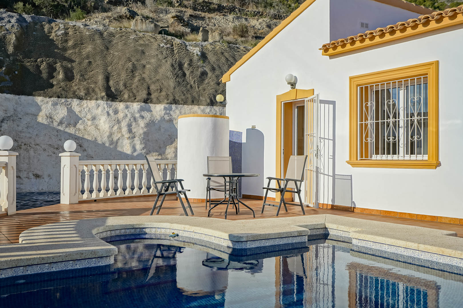 Terrasse au bord de la piscine avec mobilier de jardin, douche extérieure cylindrique et façade blanche aux encadrements ocres sur fond de colline rocheuse.