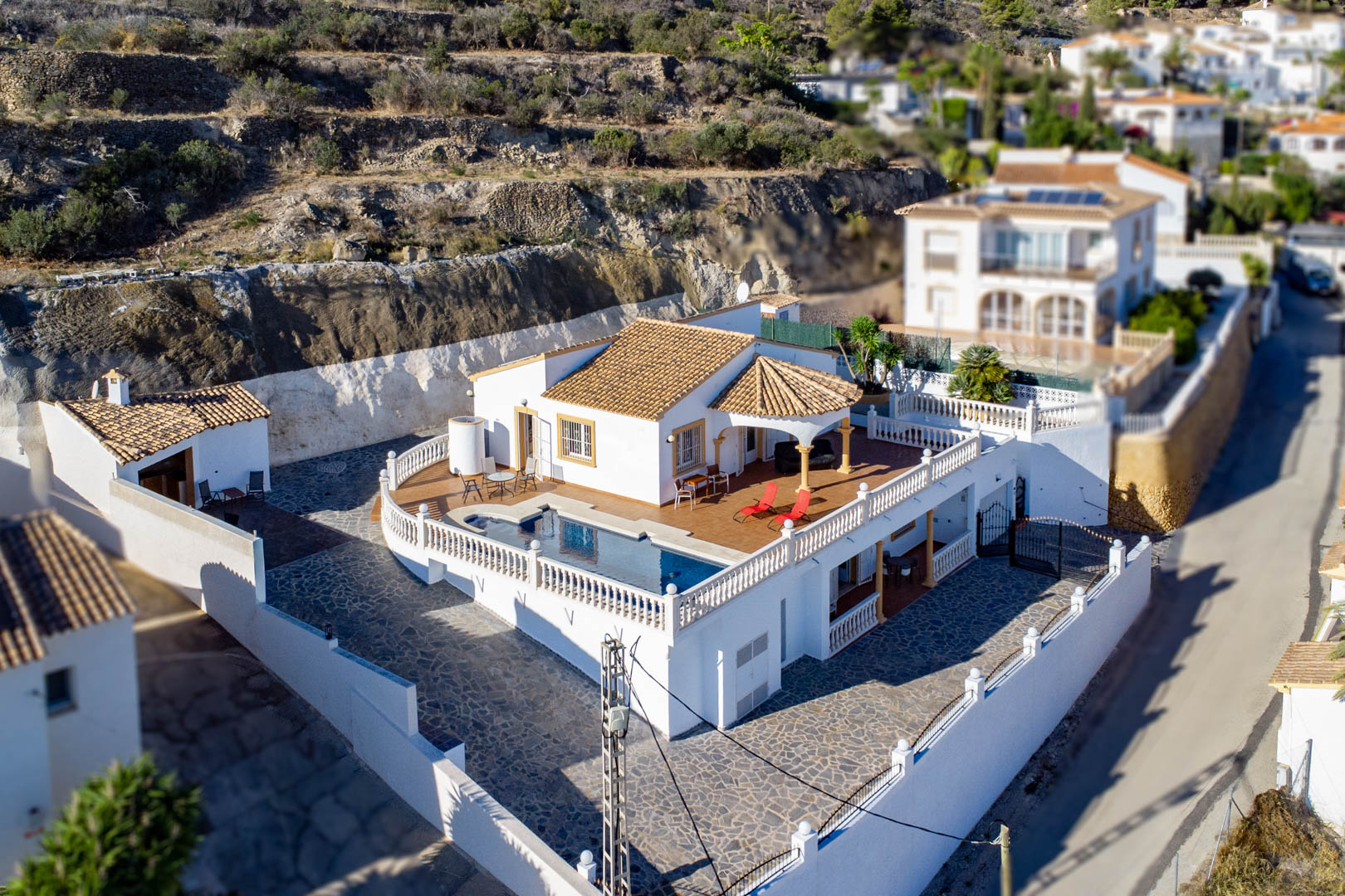 Vue aérienne de la villa avec piscine privée sur terrasse surélevée, balustrades blanches, toiture en tuiles et vaste cour pavée de pierres au pied de la colline.