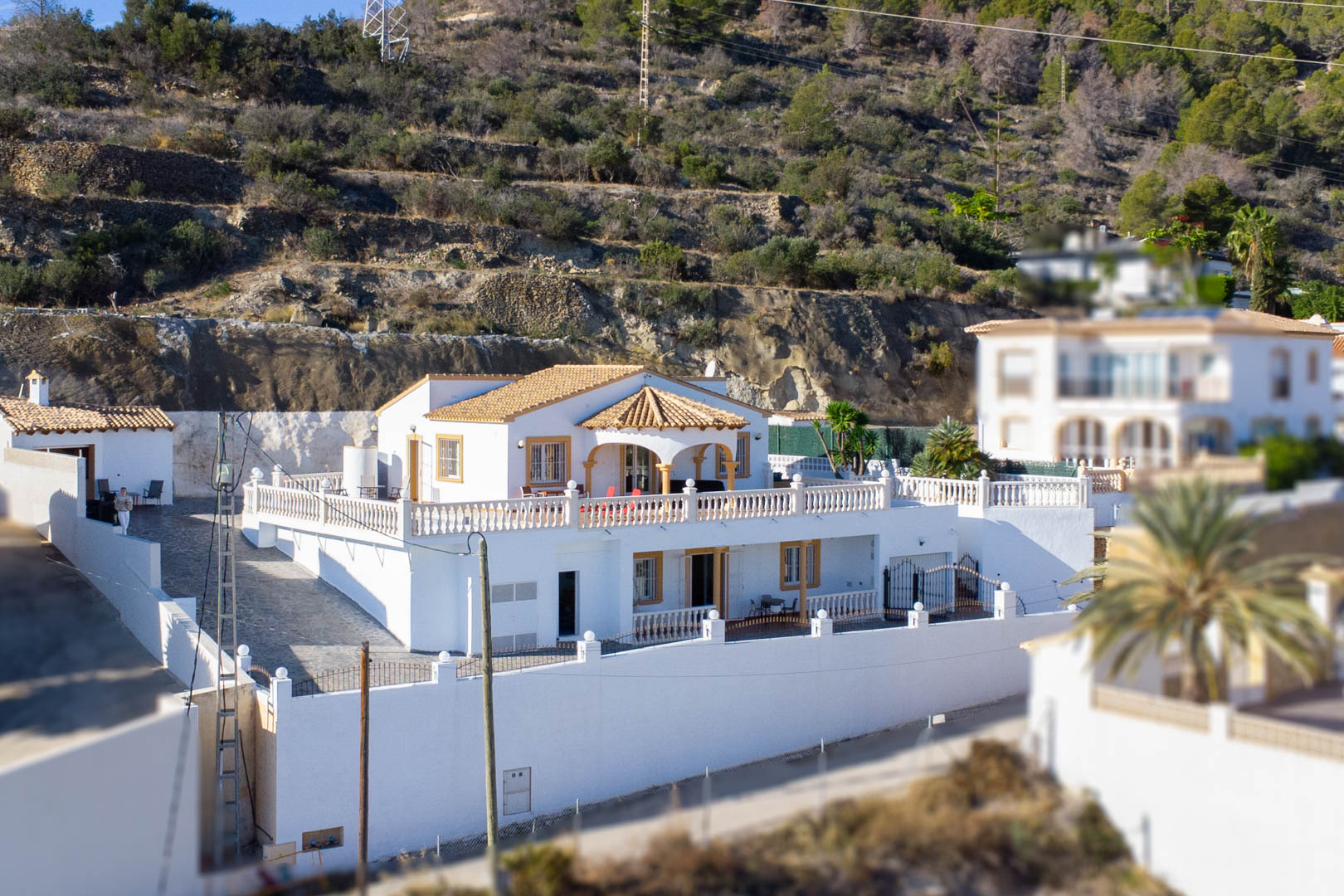 Vue extérieure de la villa sur deux niveaux avec façade blanche, toit en terre cuite et vastes terrasses à balustrades situées sur le flanc de la Sierra de Oltà.