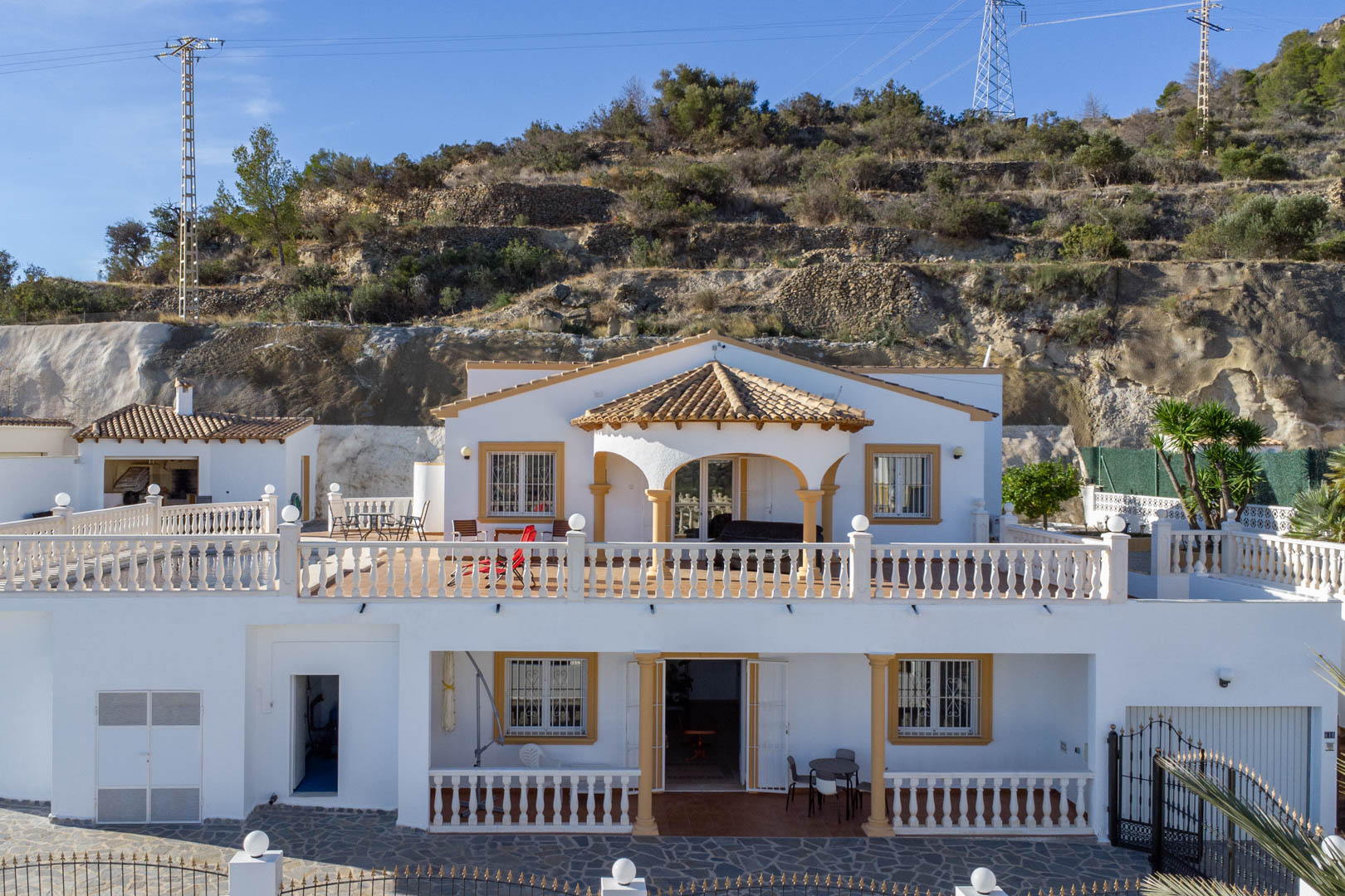 Vue extérieure de la villa sur deux niveaux avec façades blanches, balustrades classiques et porches couverts, adossée à la colline de la Sierra de Oltà.