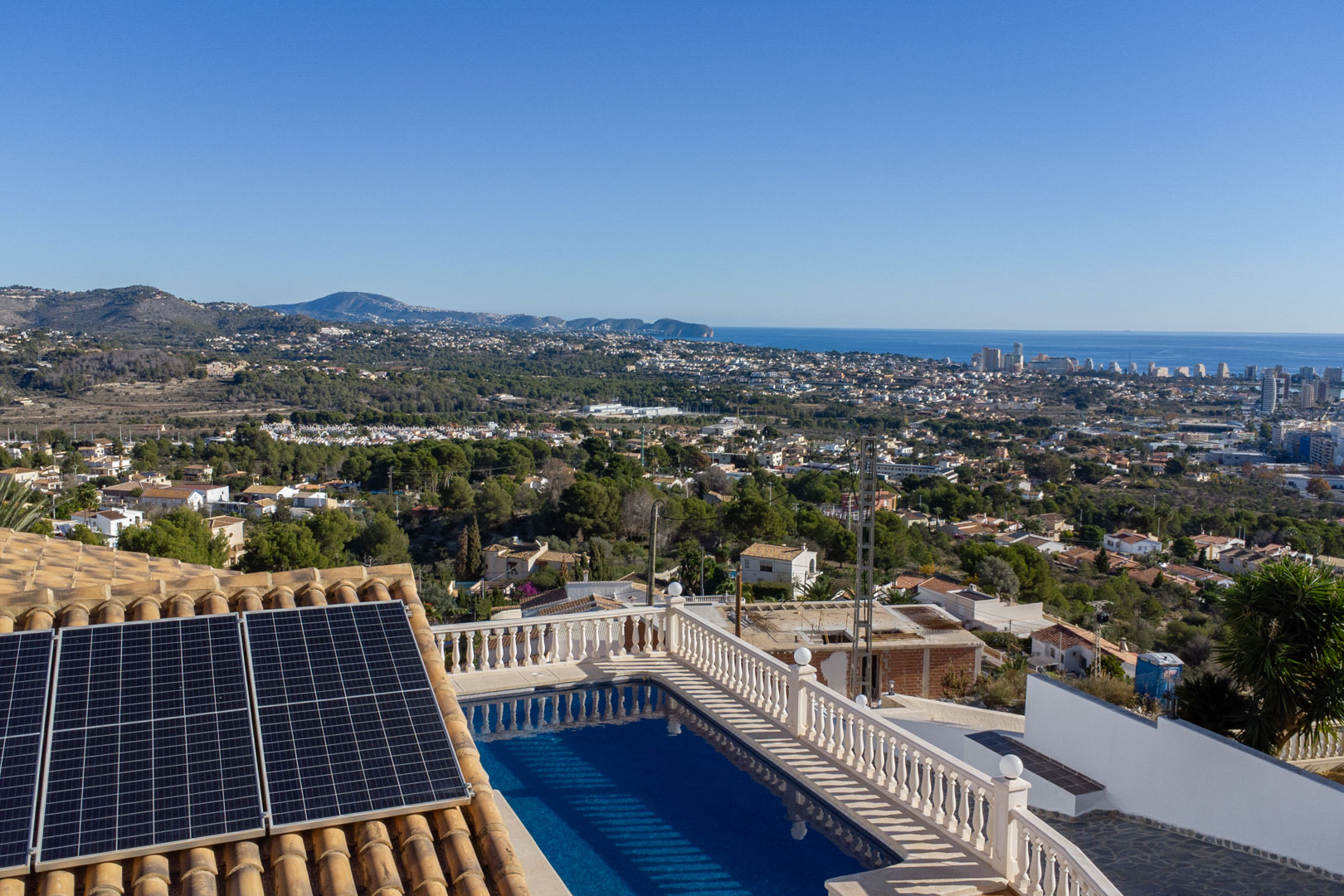 Vue surélevée montrant la piscine avec balustrade, des panneaux solaires sur le toit en tuiles et un panorama dégagé sur la côte de Calpe et la mer.