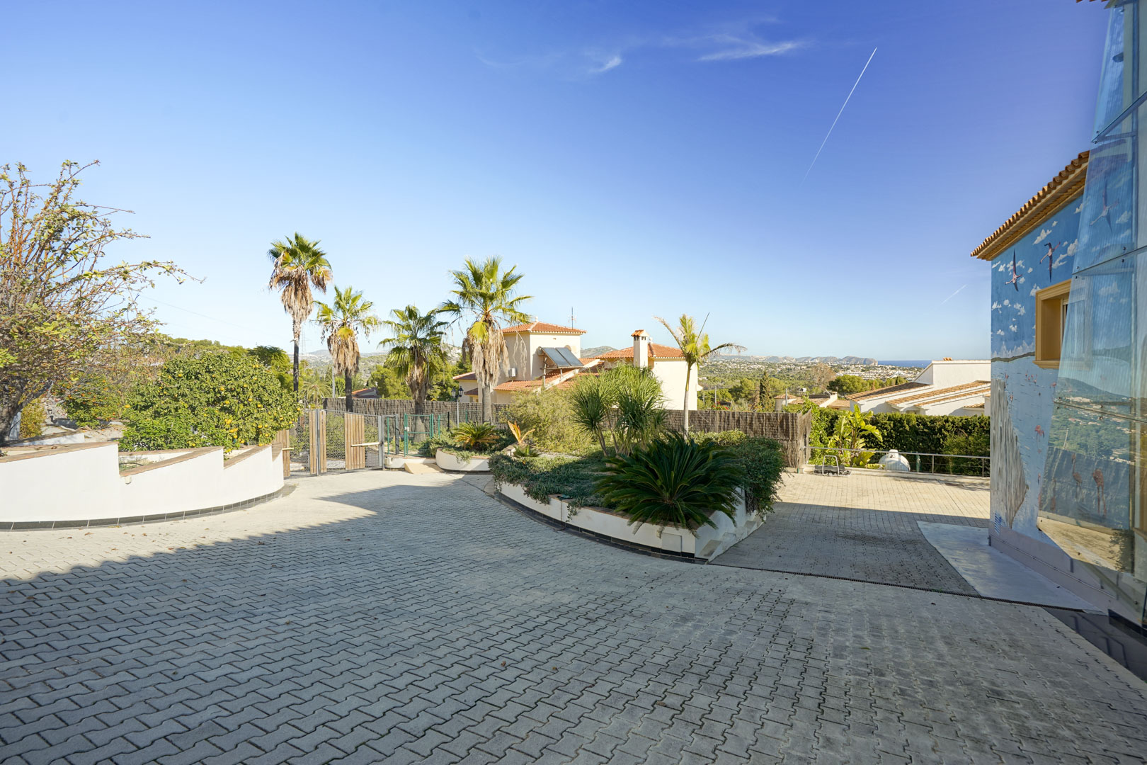 Allée d'entrée pavée avec îlot de jardin paysager, palmiers et une fresque murale décorative, offrant une vue dégagée sur les environs et la mer au loin.